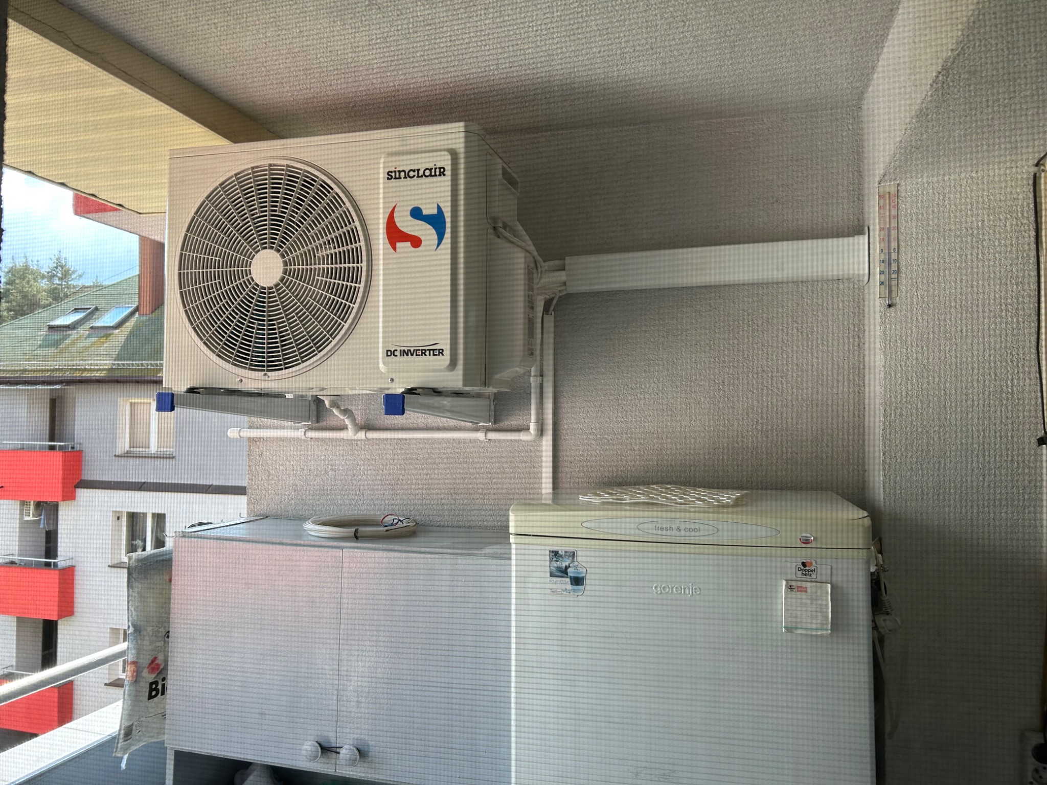 Biały klimatyzator Sinclair zamontowany na balkonie, widoczne rury instalacyjne i termometr na ścianie. W tle zabudowa balkonu i budynek mieszkalny.