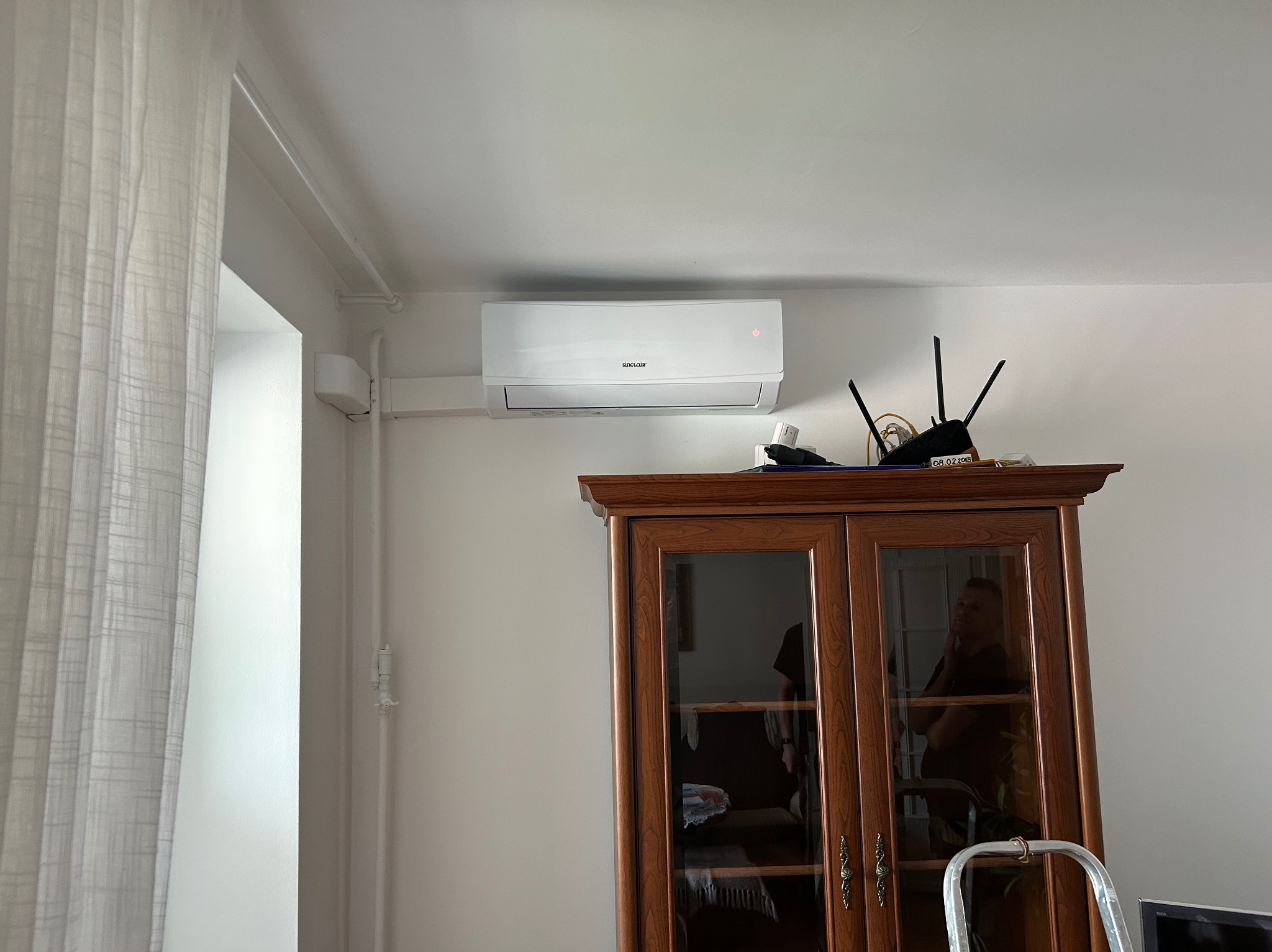 Biały klimatyzator Sinclair zamontowany nad drewnianym kredensem w jasnym wnętrzu. Widoczny fragment okna z firanką i router na szafce.