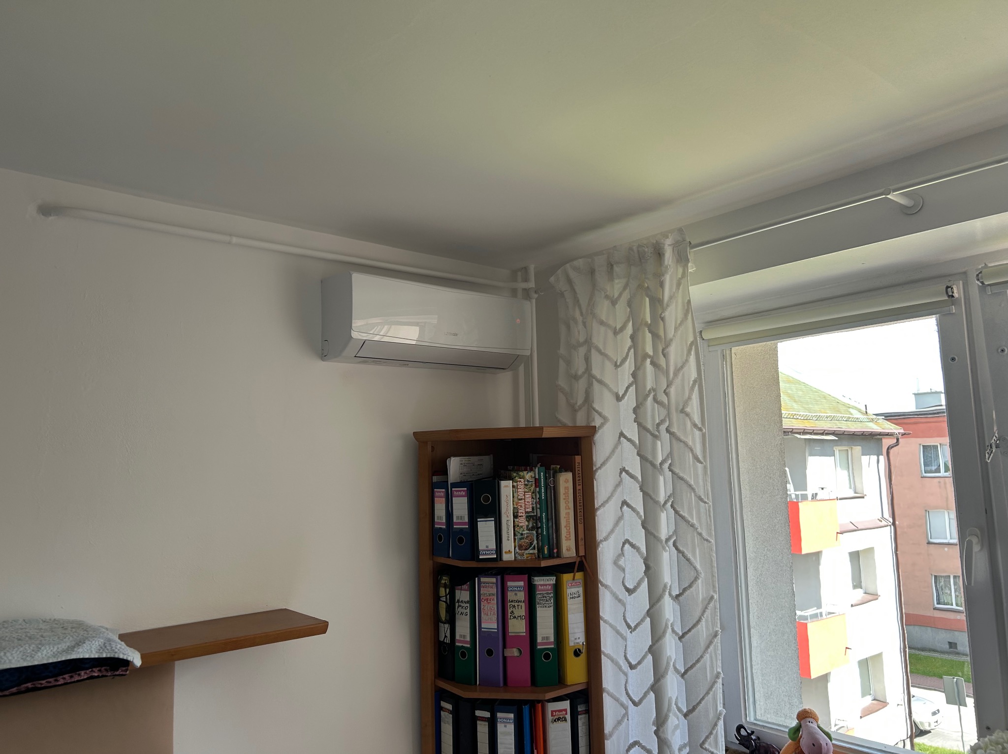 Biała klimatyzacja ścienna w pokoju z regałem na segregatory i widokiem z okna na budynek mieszkalny. Montaż klimatyzacji w mieszkaniu.
