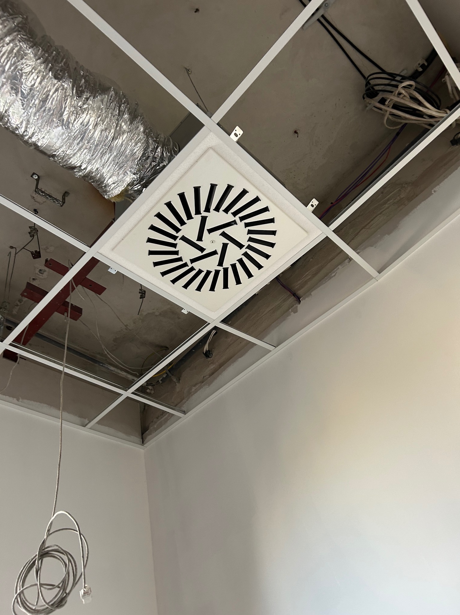 Widok z dołu na sufit podwieszany z kratką wentylacyjną o geometrycznym wzorze i aluminiowym przewodem wentylacyjnym. Widoczne kable i elementy konstrukcyjne.