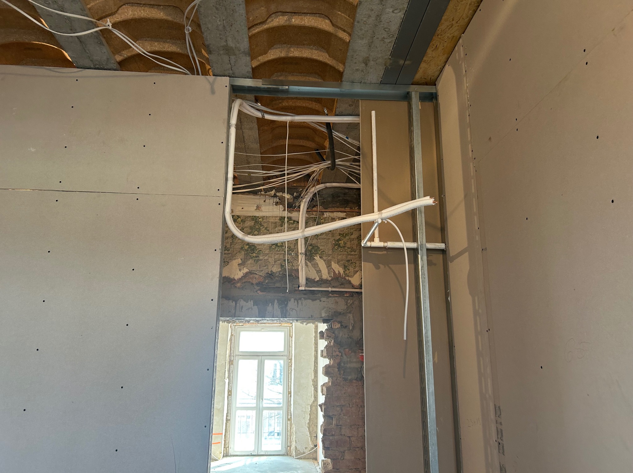 Instalacja przewodów wentylacyjnych i elektrycznych na suficie podwieszanym z płyt gipsowo-kartonowych w trakcie remontu. Widok na okno w tle.