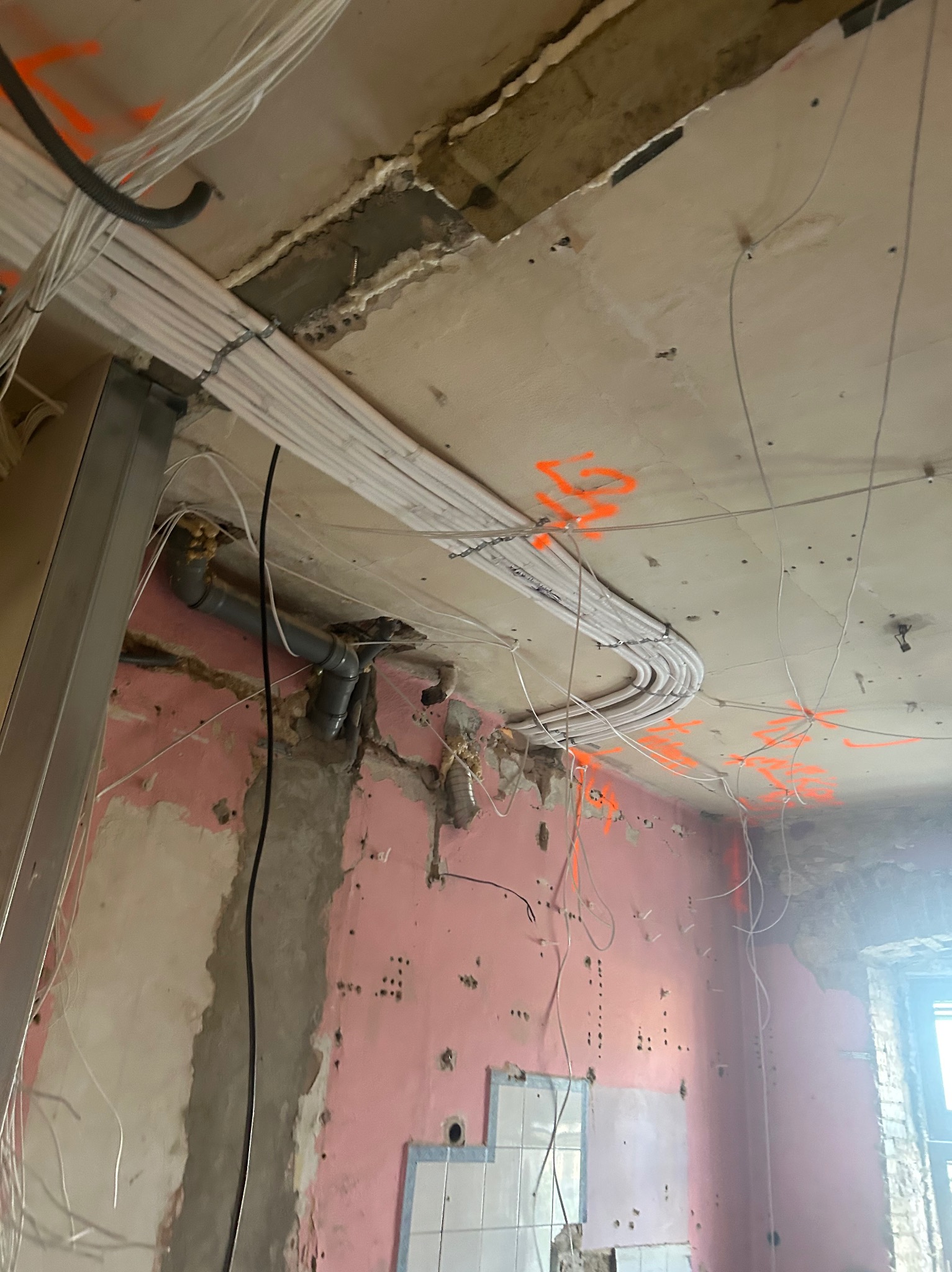 Instalacja przewodów wentylacyjnych i elektrycznych w trakcie remontu. Widoczne rury wentylacyjne pod sufitem z pomarańczowymi oznaczeniami oraz instalacja elektryczna.