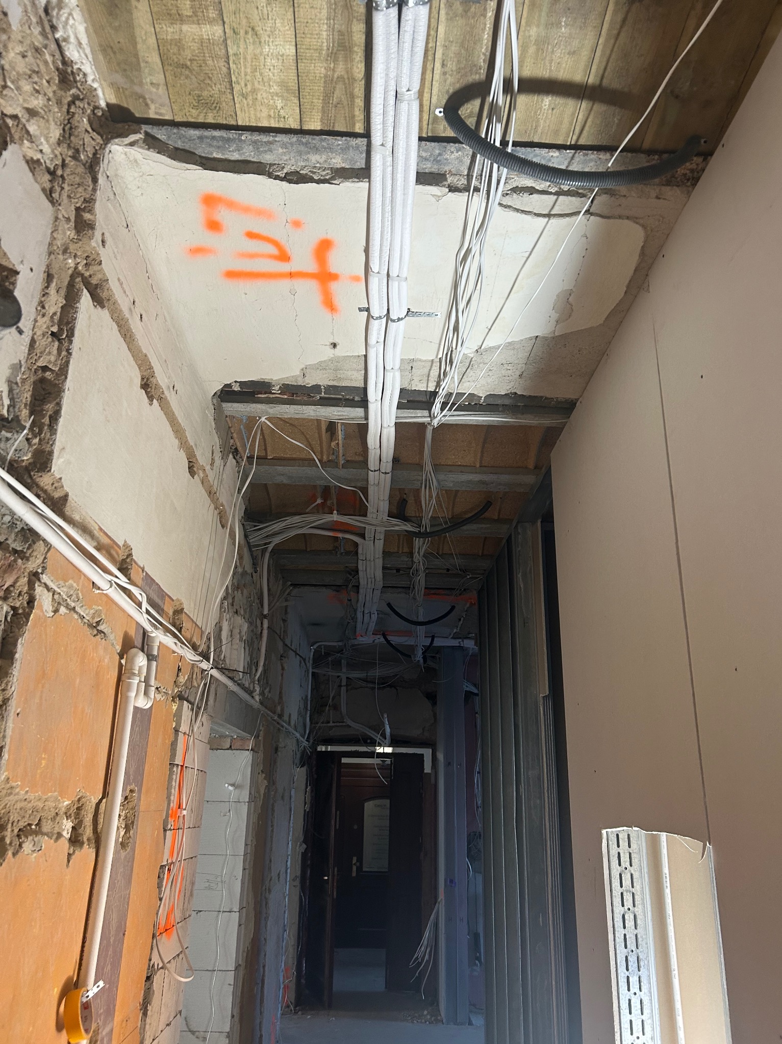Montaż instalacji w surowym wnętrzu: białe rury i kable prowadzone pod drewnianym stropem, ściany w trakcie wykończenia z pomarańczowymi oznaczeniami, widok w głąb korytarza.