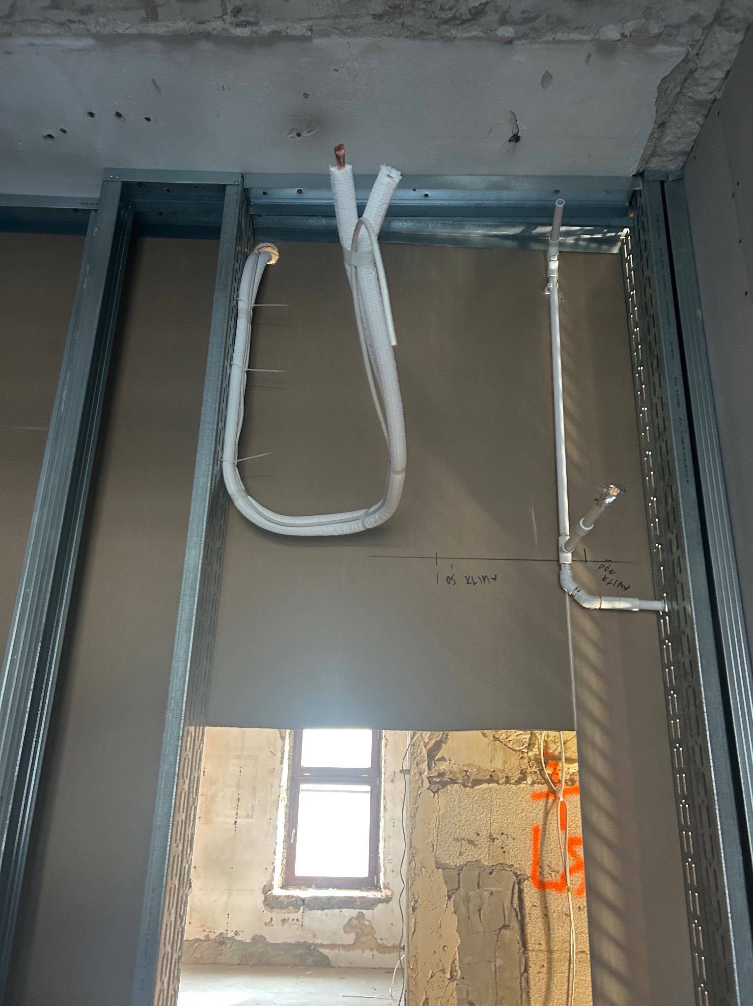 Instalacja przewodów klimatyzacji w budynku w trakcie remontu, widoczne profile metalowe i ściana z instalacją. Przewody białe, częściowo w izolacji. Widok przez otwór w ścianie.