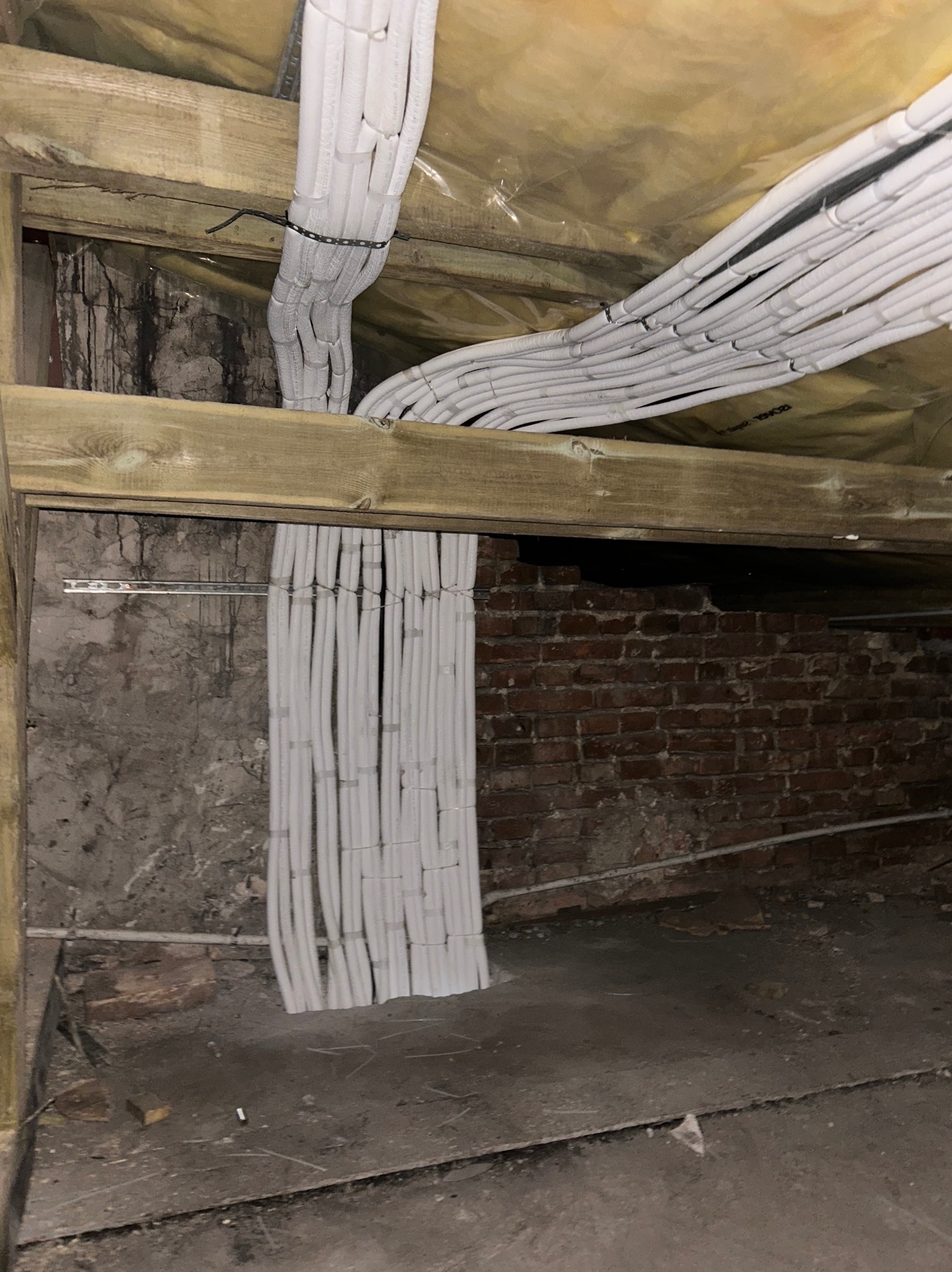 Równolegle ułożone białe rury wentylacyjne pod drewnianą konstrukcją stropu, z widoczną ścianą z cegły i elementami instalacji. Niska ekspozycja, surowy stan wykończenia.