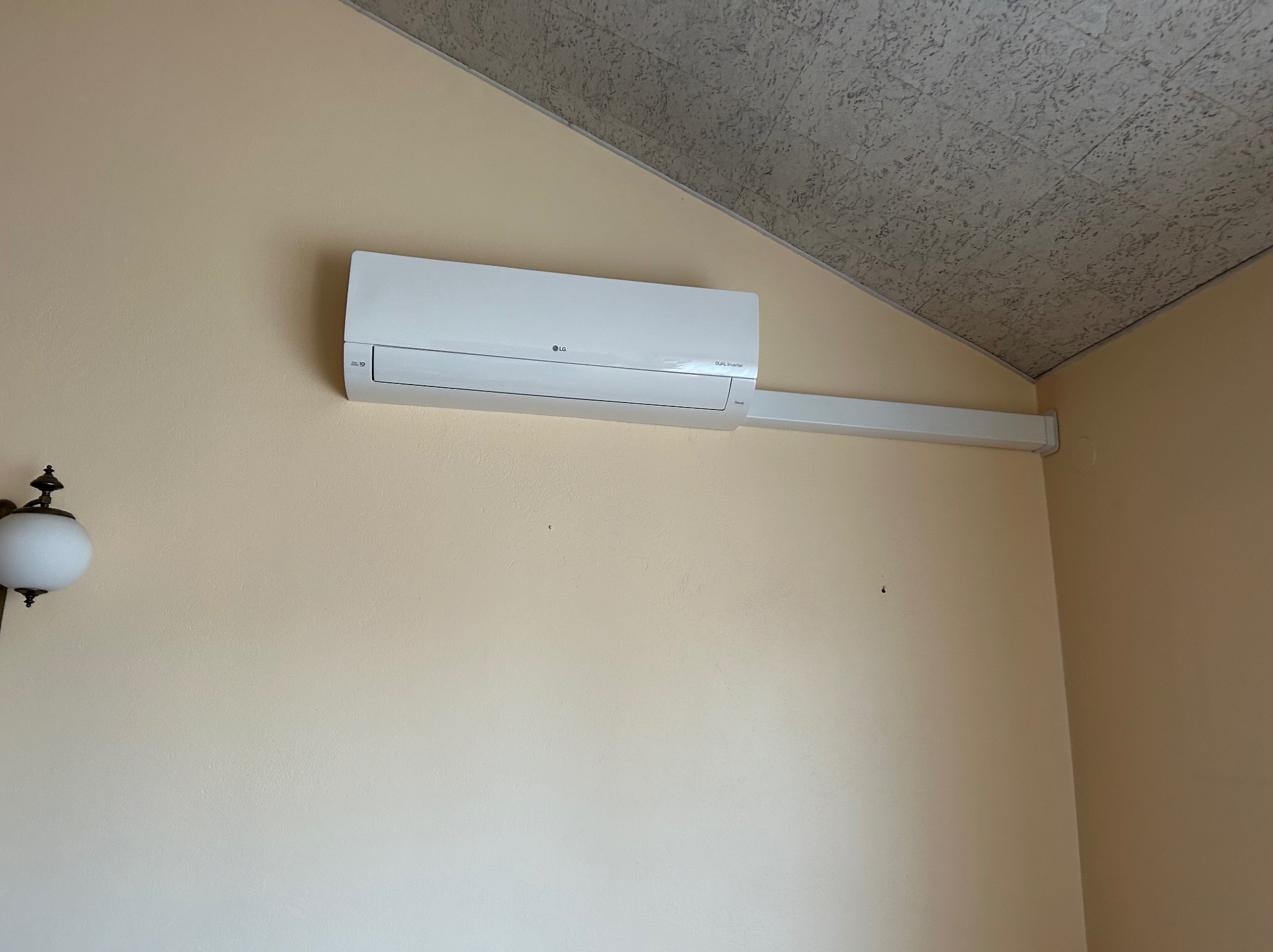 Biała klimatyzacja LG Dual Inverter zamontowana na beżowej ścianie pod skośnym sufitem. Widoczna listwa maskująca instalację. Lampa w lewym dolnym rogu.