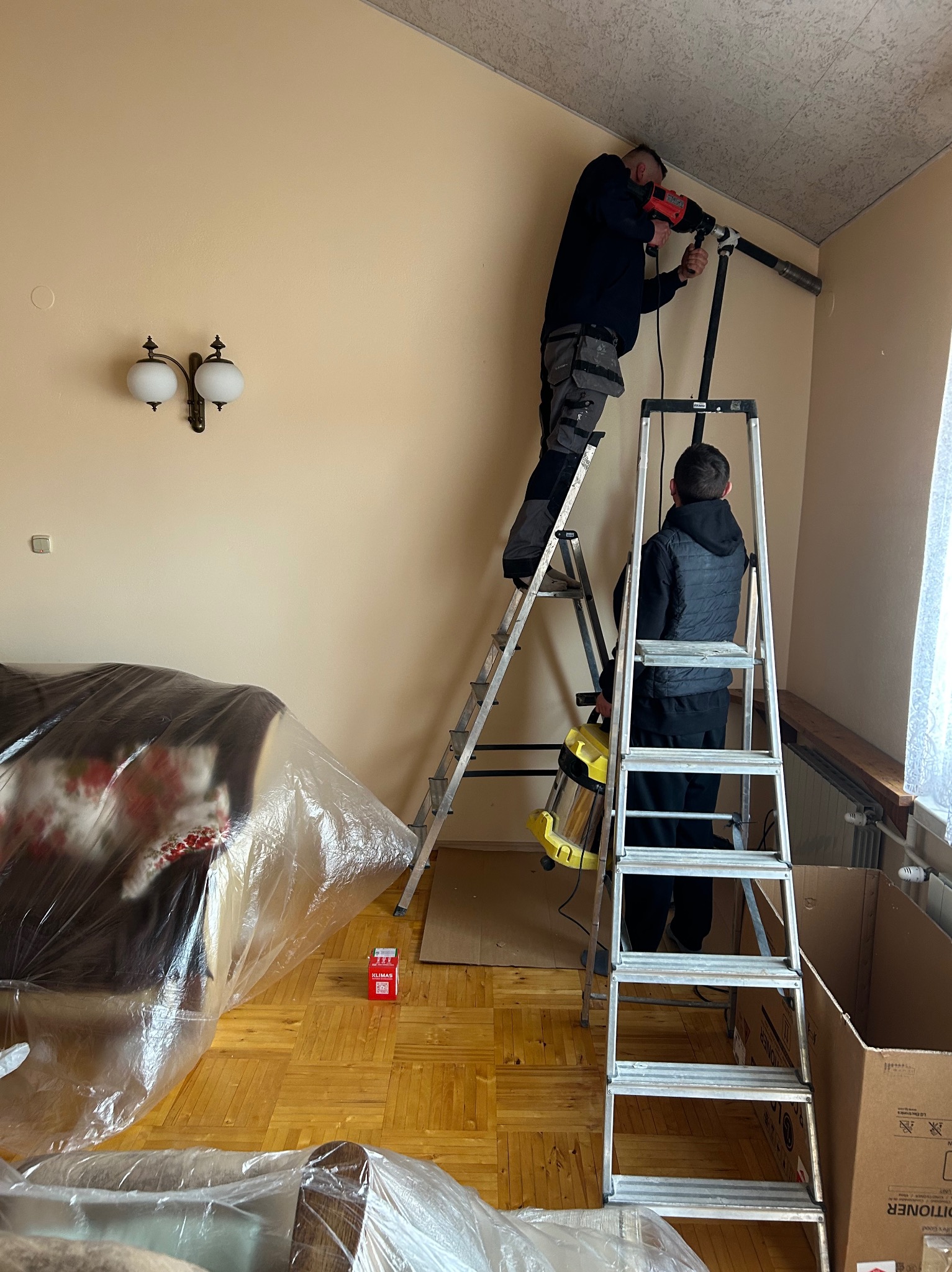 Montaż klimatyzacji: Dwóch monterów na drabinach wierci otwór w ścianie, zabezpieczone meble folią, widoczna lampa robocza i kartonowe pudła.