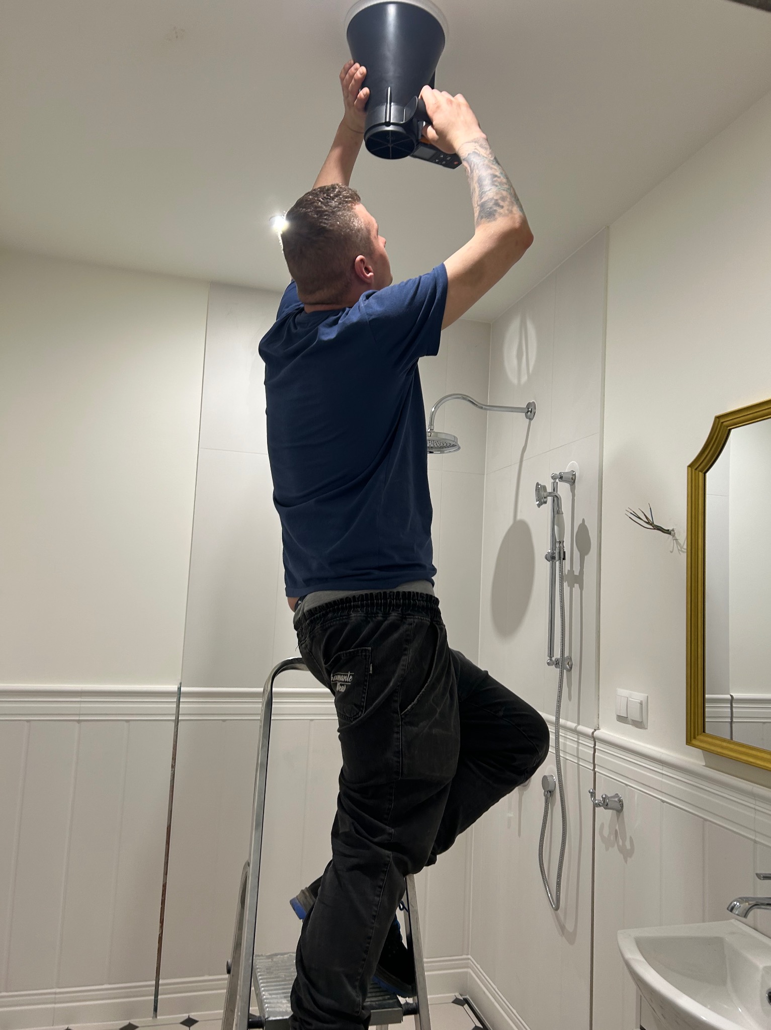 Instalator na drabinie montuje anemostat sufitowy w łazience z białą armaturą i włączonym oświetleniem. Widoczne przewody wentylacyjne.