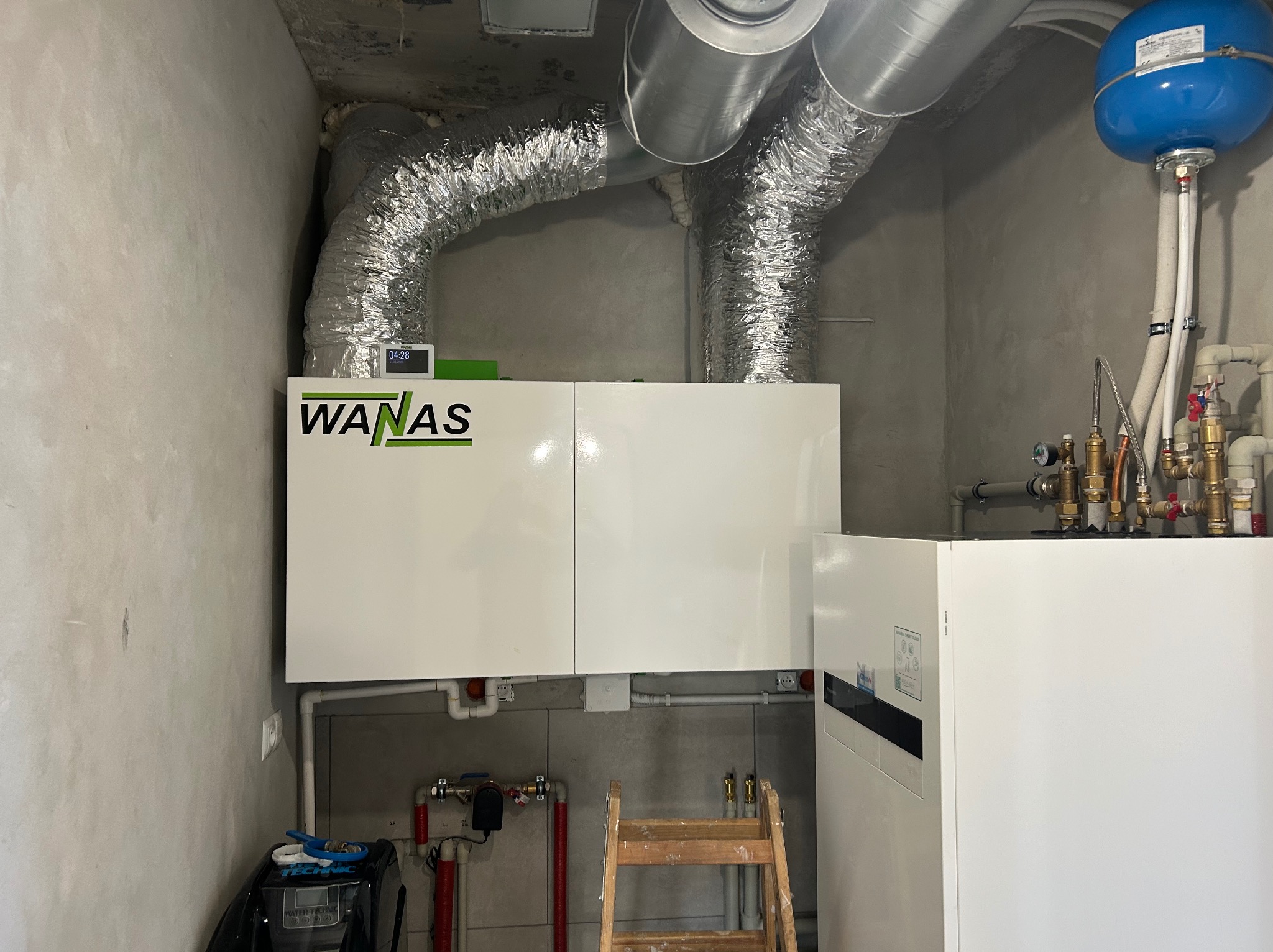 Zmontowana instalacja rekuperacji WANAS z aluminiowymi przewodami wentylacyjnymi w szarej kotłowni. Widoczny niebieski zbiornik wyrównawczy i drabina.