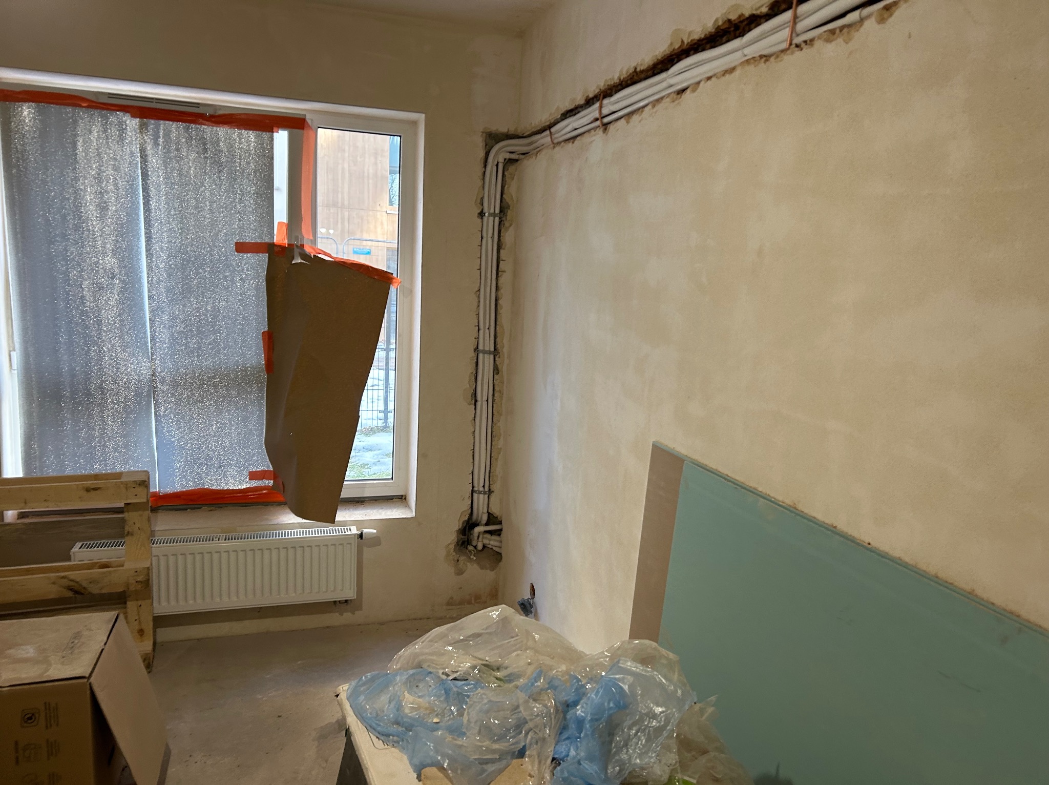 Instalacja przewodów wentylacyjnych w bruzdach ściany, zakryte okno folią ochronną podczas remontu. Widoczne materiały budowlane i grzejnik.
