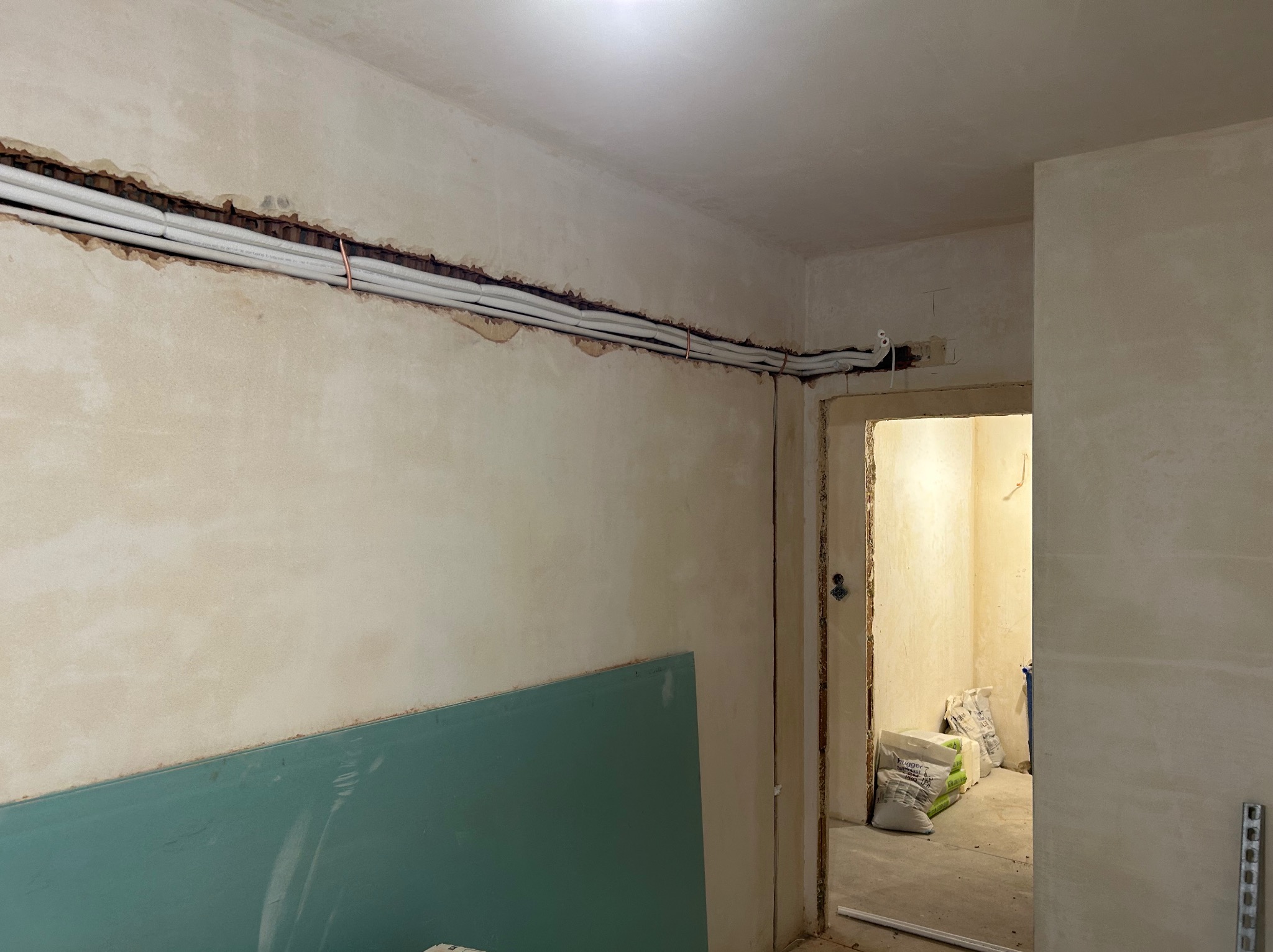 Montaż instalacji wentylacyjnej w Zrębice: Białe rury prowadzone w wyżłobieniu ściany, przygotowane do zakrycia. Widoczne materiały budowlane i płyta gipsowa.