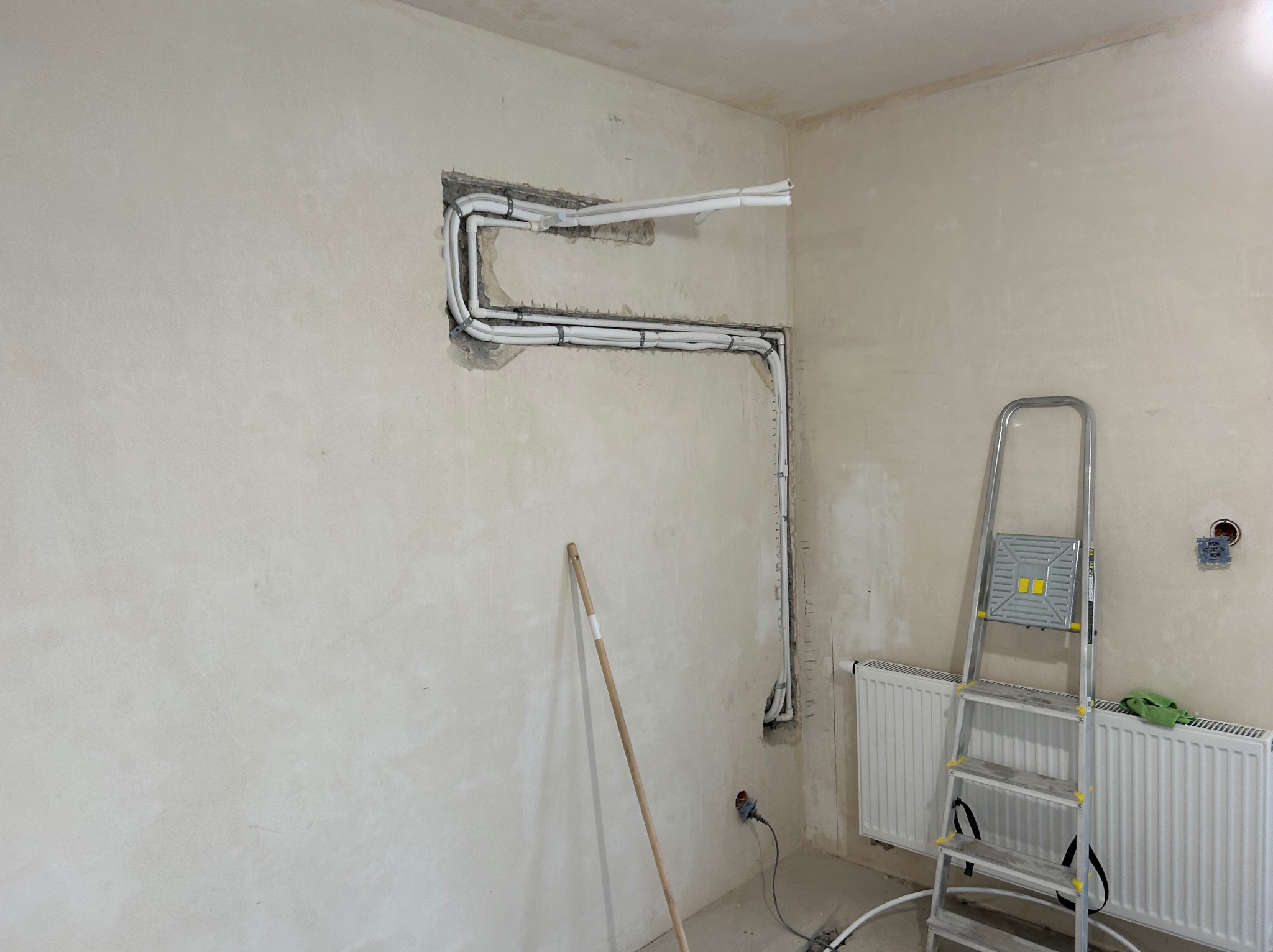 Montaż instalacji wentylacyjnej z białych rur w ścianie podczas remontu, widoczna drabina i narzędzia. Surowy stan ścian.