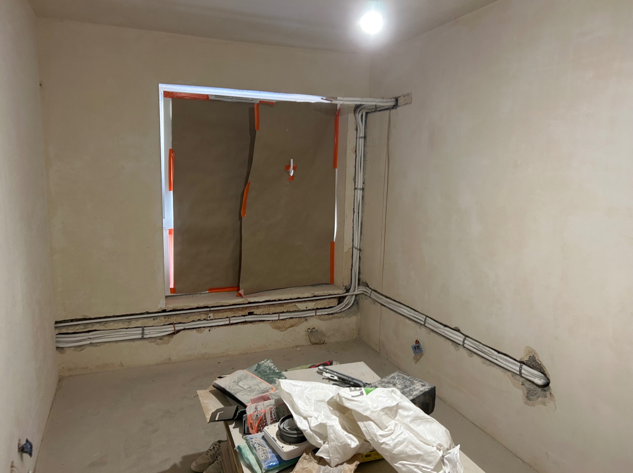Wnętrze w trakcie prac instalacyjnych: widoczne kable poprowadzone wzdłuż ścian, okno zabezpieczone kartonem, narzędzia na podłodze. Stan surowy, przygotowanie do wykończenia.