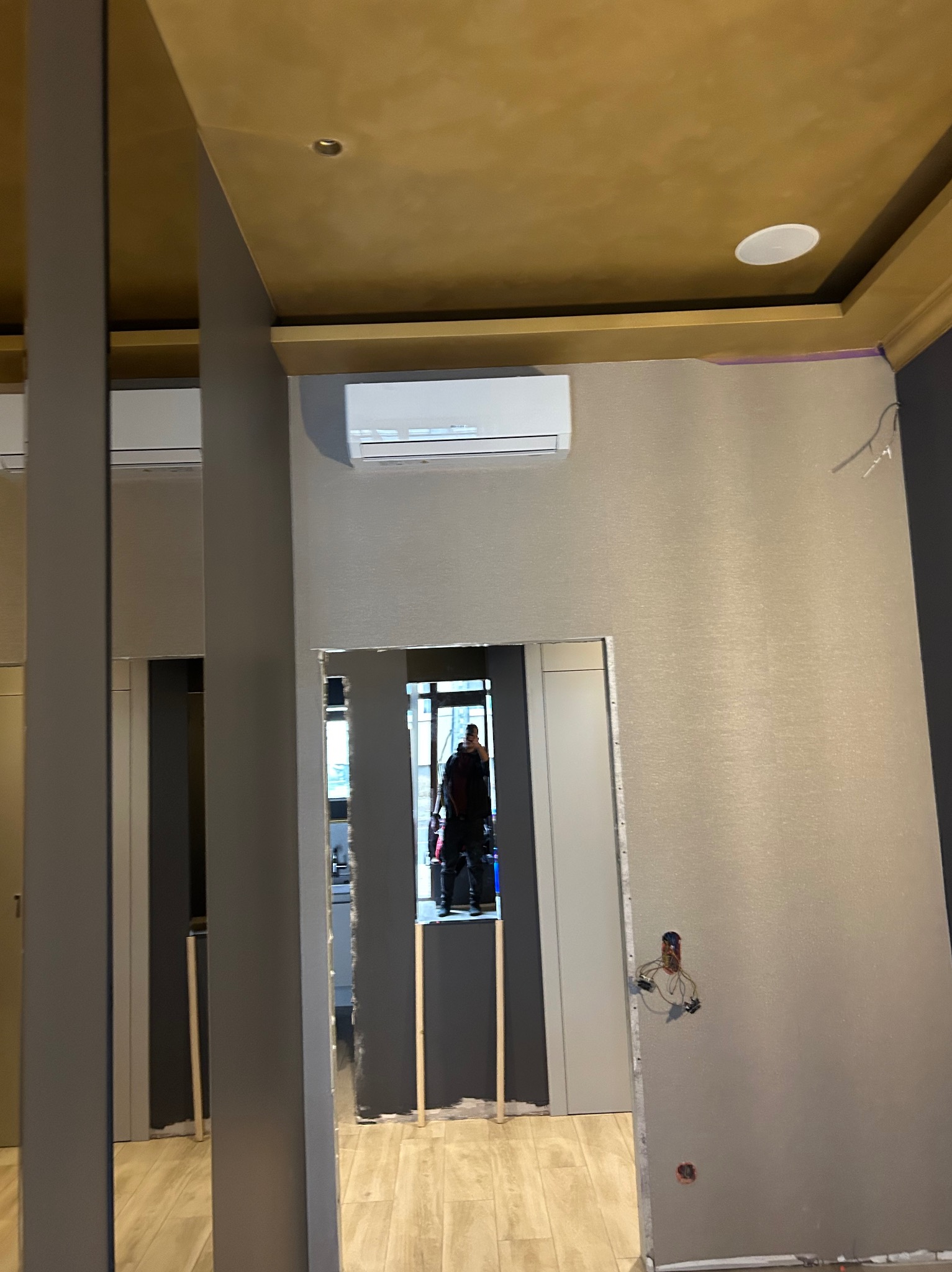 Montaż klimatyzacji split w pomieszczeniu z szarą tapetą i złotym sufitem. Widoczne przewody i otwory w ścianie przed wykończeniem.