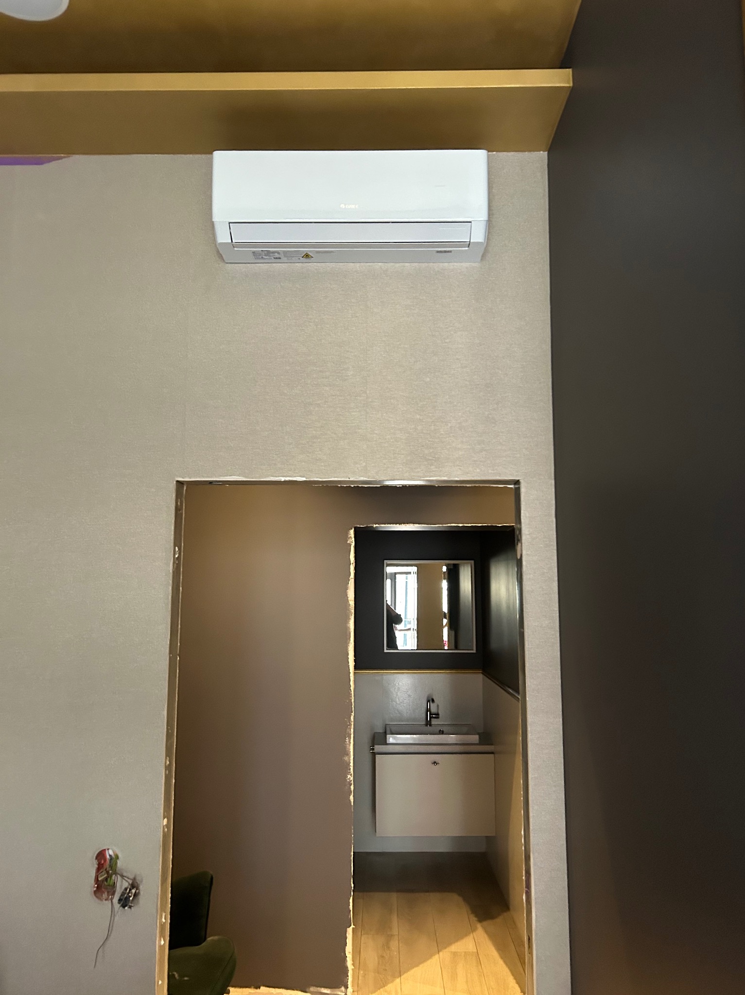 Biała klimatyzacja na ścianie nad wejściem do łazienki z widoczną umywalką i lustrem. Beżowa tapeta i ciemnoszara ściana. Złote wykończenia.