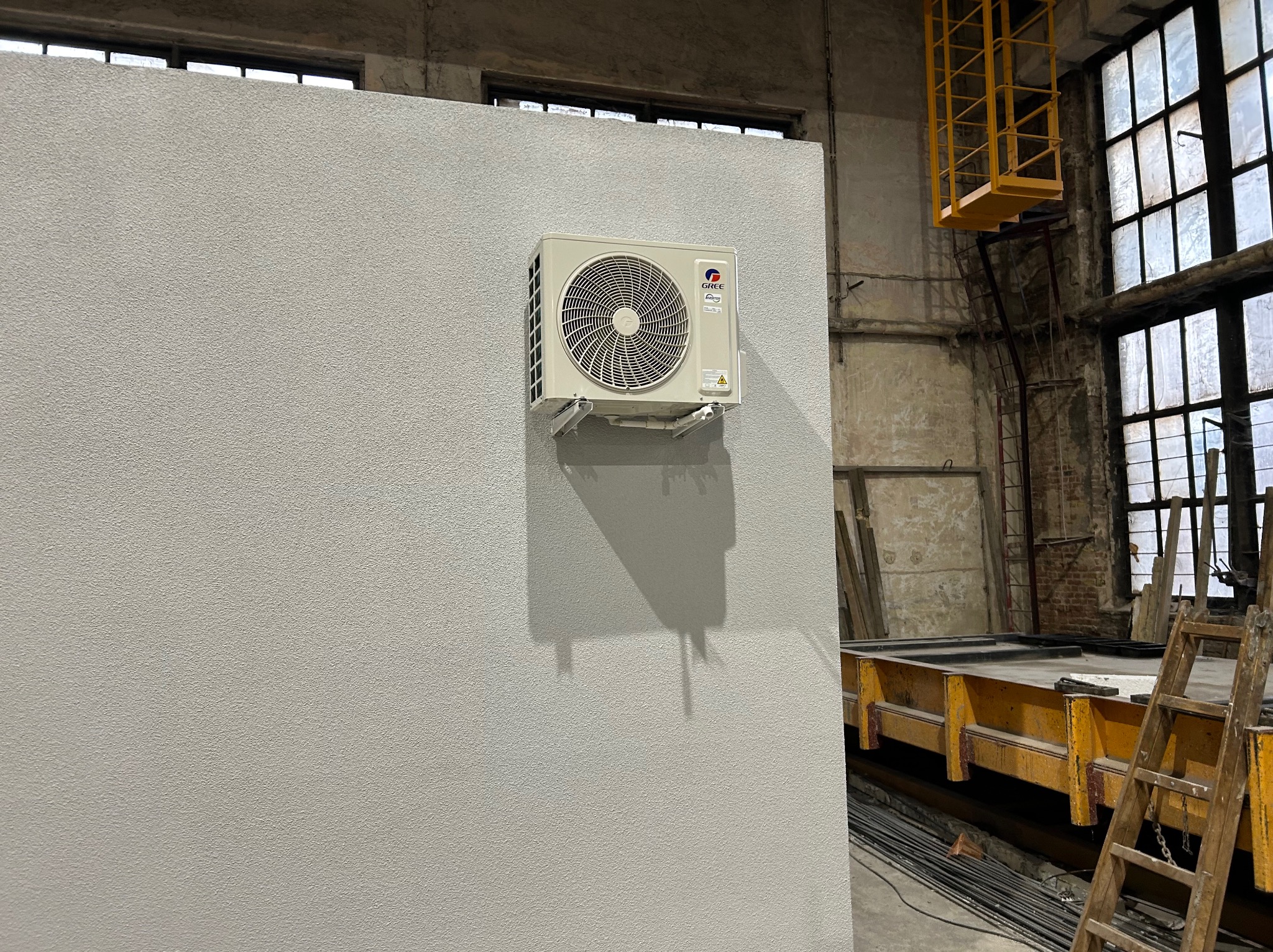 Biała jednostka klimatyzacji marki Gree zamontowana na szarej ścianie o teksturowanej powierzchni w surowym wnętrzu hali z oknami i żółtym podnośnikiem.