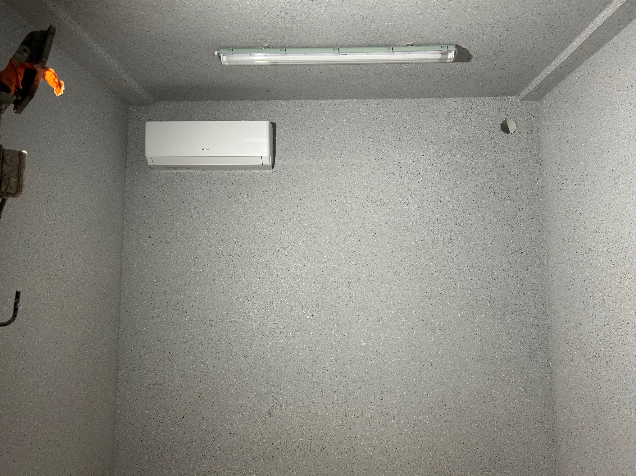 Zamontowany, biały klimatyzator na szarej, teksturowanej ścianie. Nad nim podłużna lampa. W rogu widoczne narzędzia.