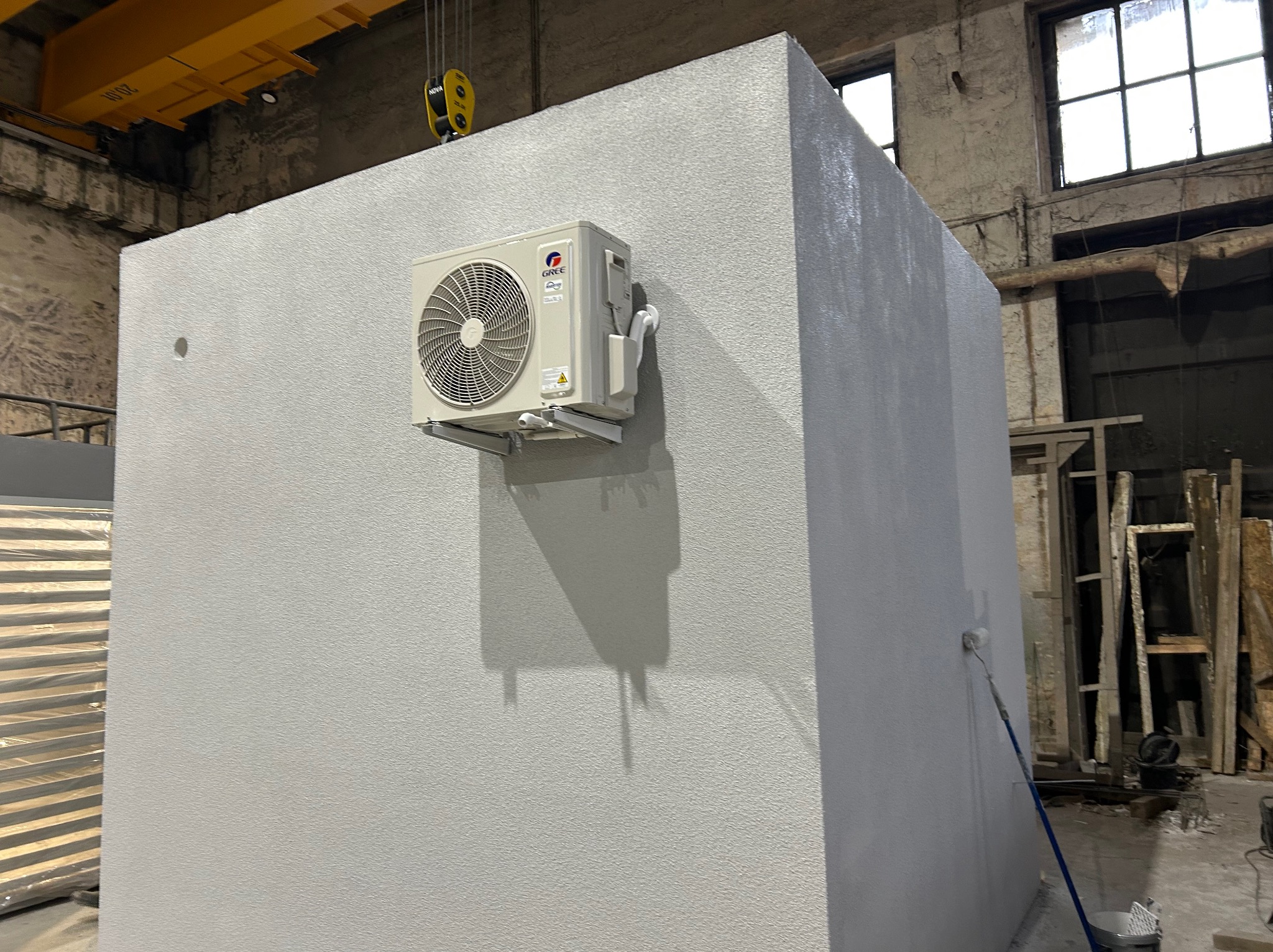 Biały klimatyzator Gree zamontowany na ścianie w surowym wnętrzu przemysłowym, widoczne okna i suwnica. Malowanie ściany w toku.