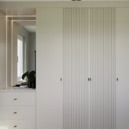 Minimalistyczna zabudowa wnęki z przesuwnymi drzwiami z pionowymi lamelami. Szafka z trzema szufladami i lustro z podświetleniem LED. Doniczka z rośliną na blacie.