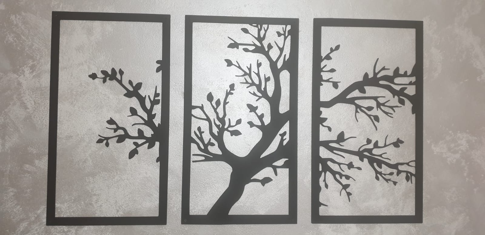 Metalowa dekoracja ścienna: trzy panele z wyciętymi laserowo sylwetkami drzew na tle teksturowanej ściany.