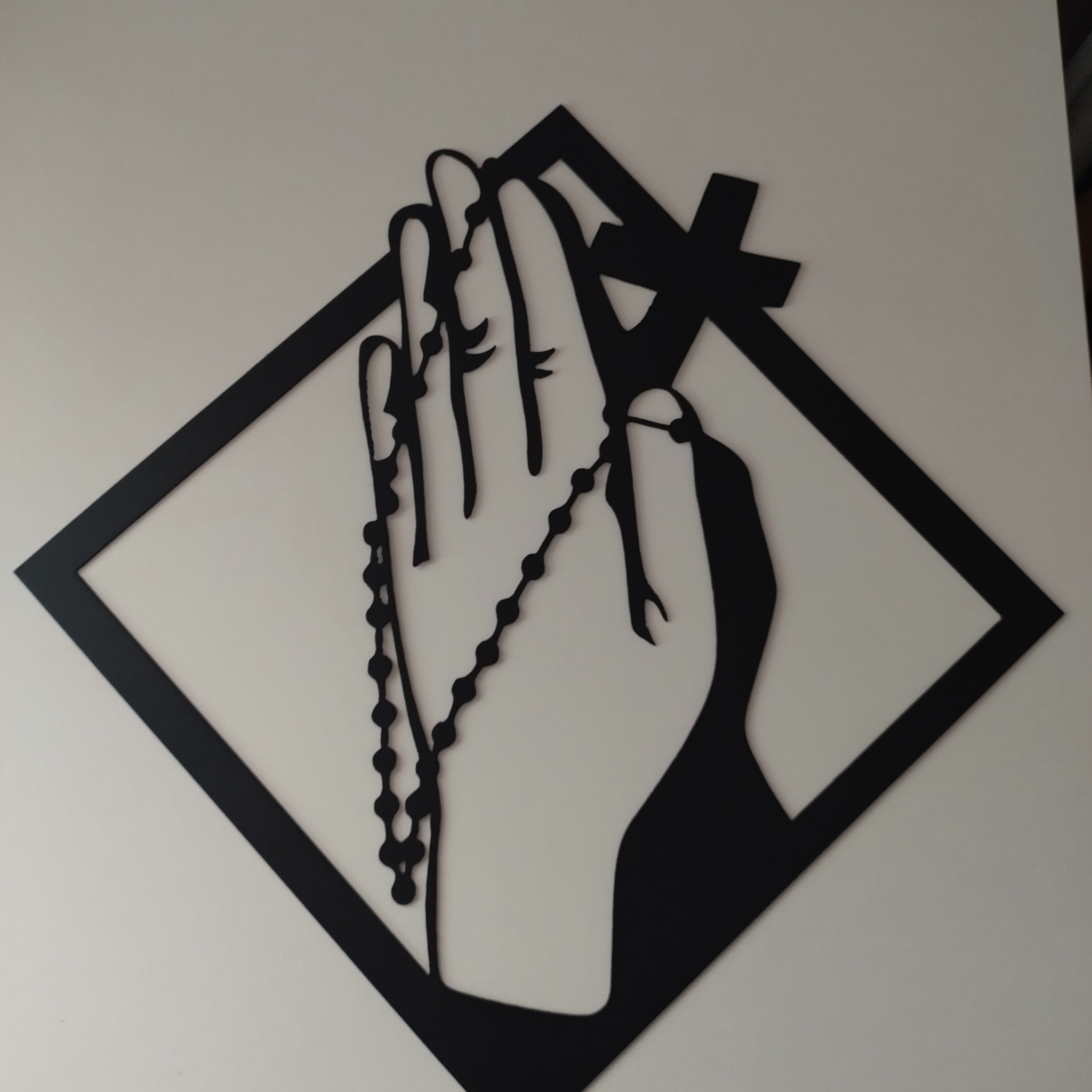 Czarna, ażurowa dekoracja ścienna przedstawiająca dłoń trzymającą różaniec, umieszczona w rombowej ramie.