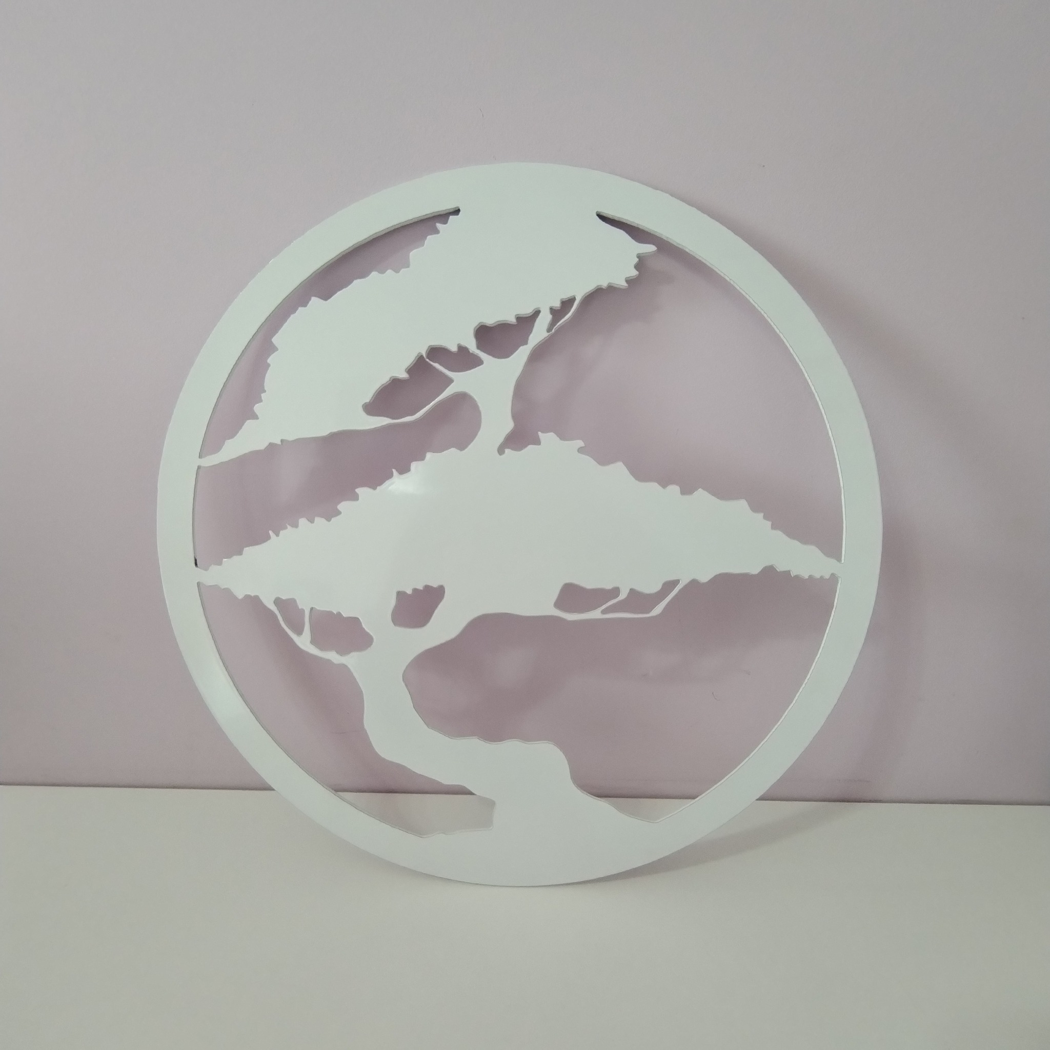 Biała, okrągła dekoracja ścienna z motywem drzewa wycięta techniką CNC, umieszczona na tle jasnoróżowej ściany i białego blatu.
