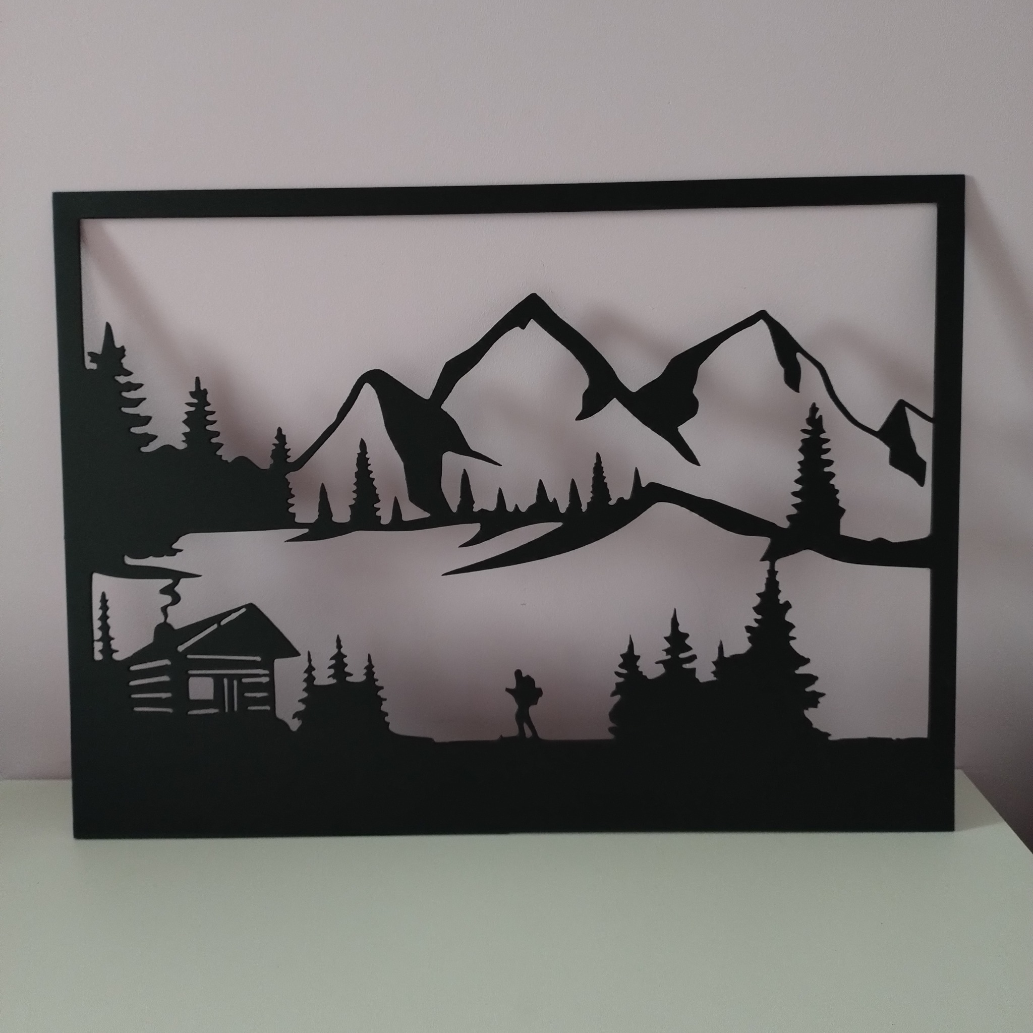 Czarna, metalowa dekoracja ścienna przedstawiająca górski krajobraz z chatą, drzewami i postacią wędrowca na tle jasnej ściany.
