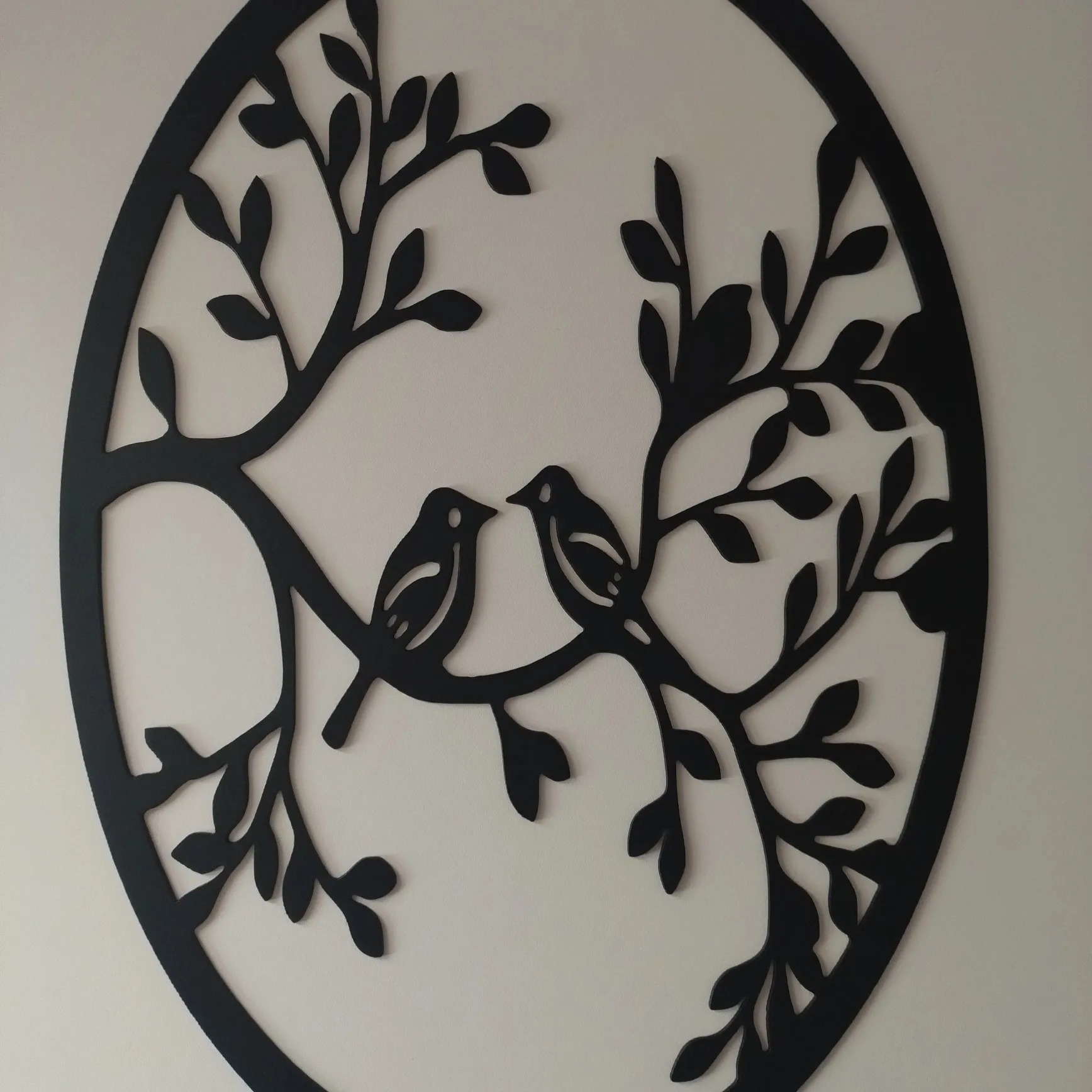 Dekoracja ścienna: czarny, ażurowy okrąg z motywem drzewa i dwóch siedzących obok siebie ptaków, na białym tle ściany.