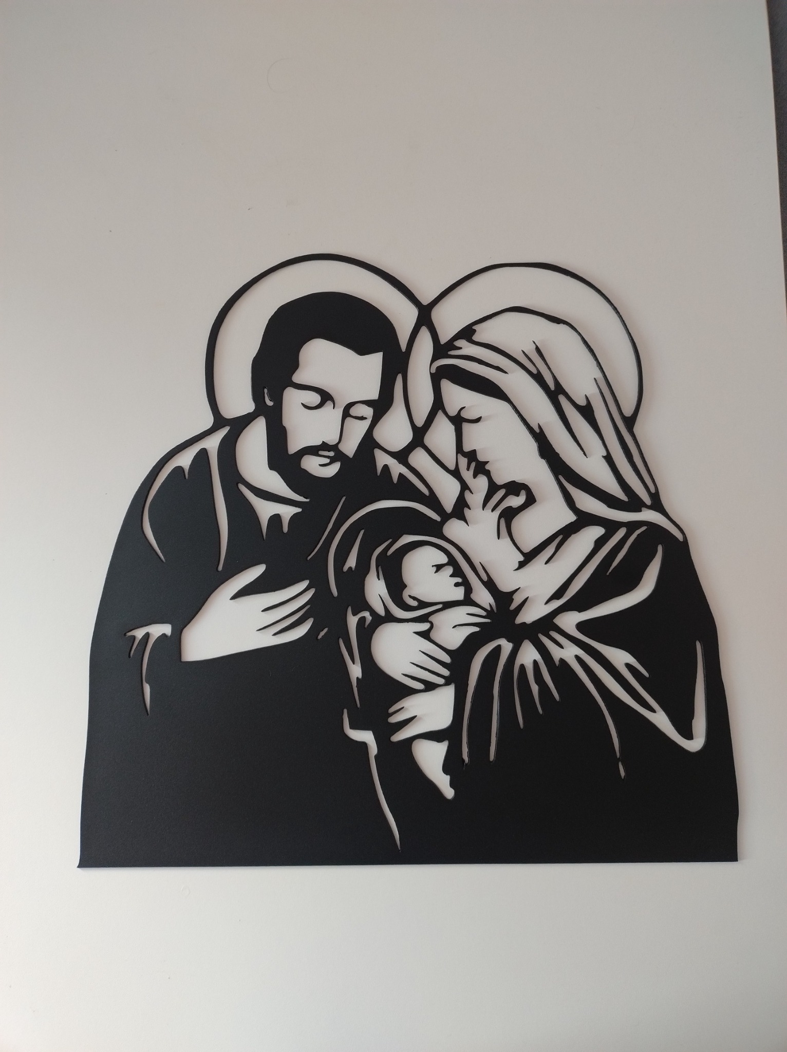 Wycięty z metalu, czarny obraz przedstawiający Świętą Rodzinę na białym tle. Widoczne delikatne, laserowo wycięte detale.
