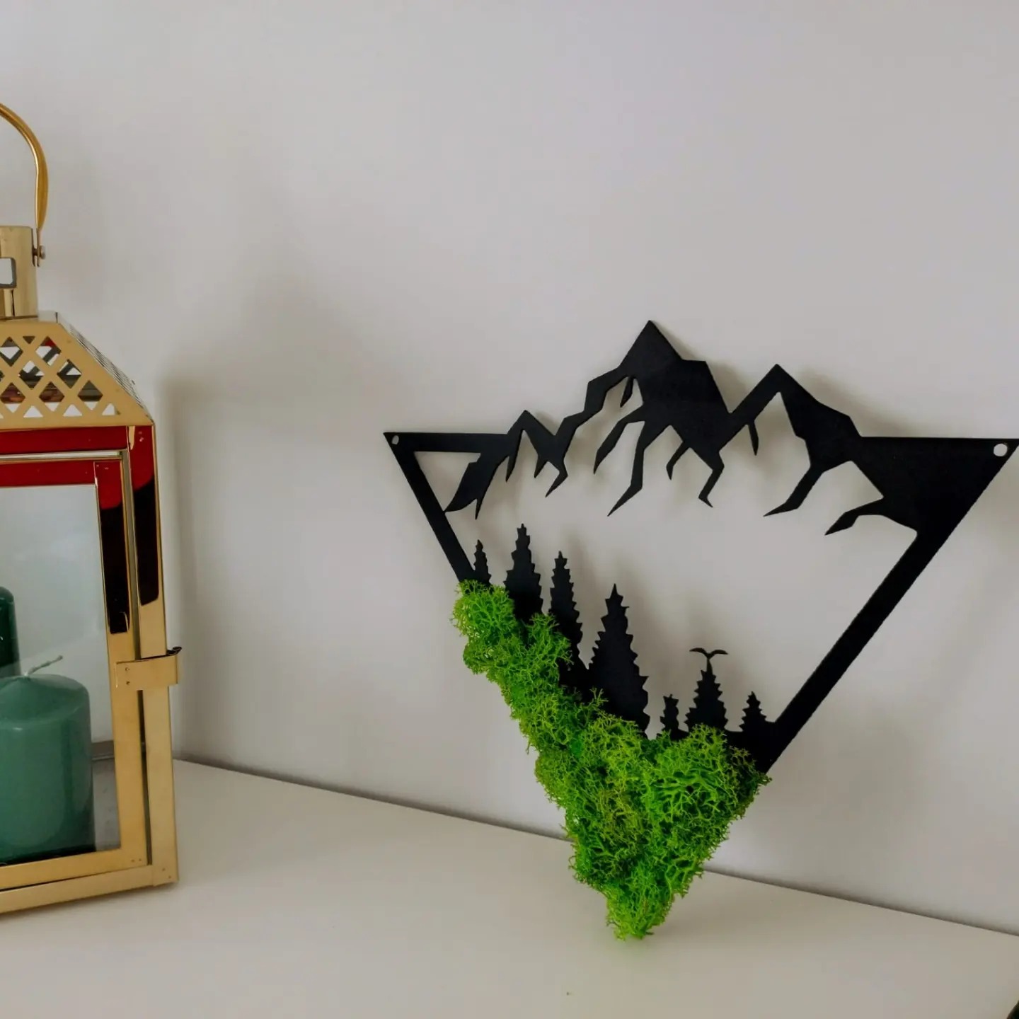 Dekoracja ścienna: czarny metalowy trójkąt z wyciętym motywem gór i lasu, wypełniona zielonym mchem, obok złotej latarenki z zielonymi świecami.