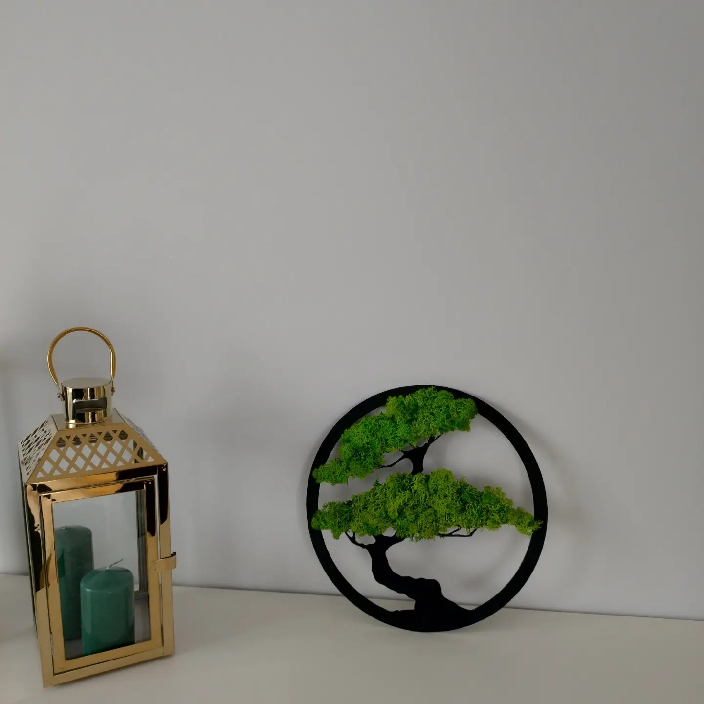 Dekoracja ścienna: czarny okrągły panel z mchem imitującym drzewko bonsai, obok złotej latarenki z zielonymi świecami, na białym tle.