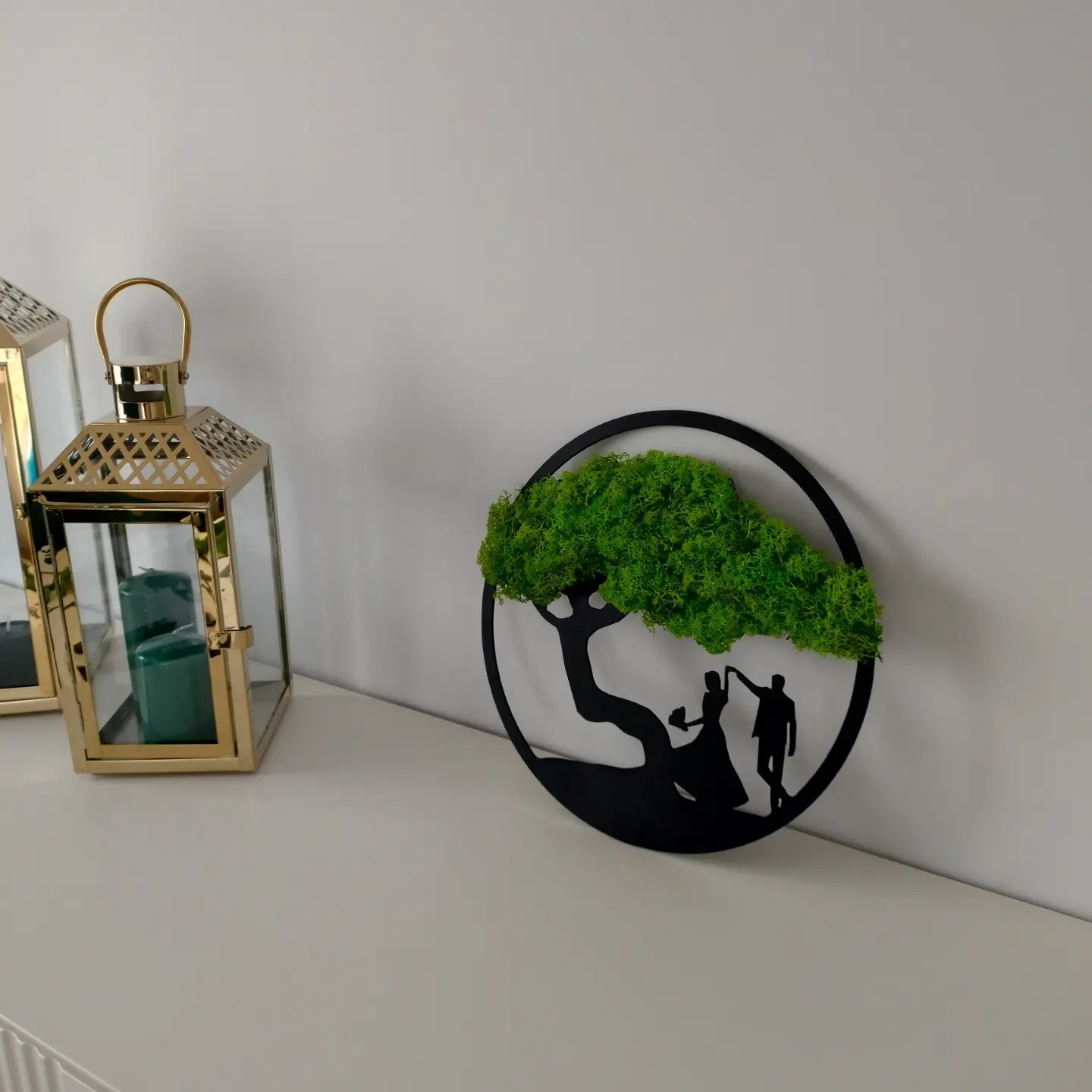Dekoracja ścienna: czarny metalowy okrąg z sylwetką pary młodej pod drzewem z mchu, obok złote lampiony ze świecami.