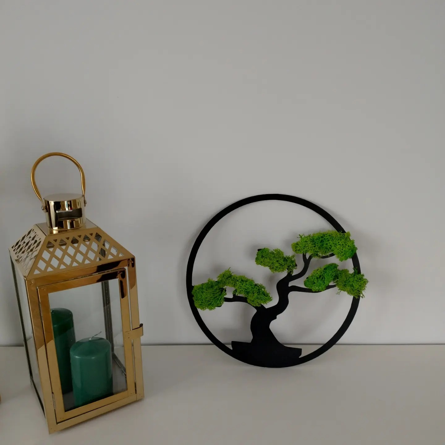 Złota lampion z zielonymi świecami obok czarnej dekoracji ściennej przedstawiającej drzewo z mchu.