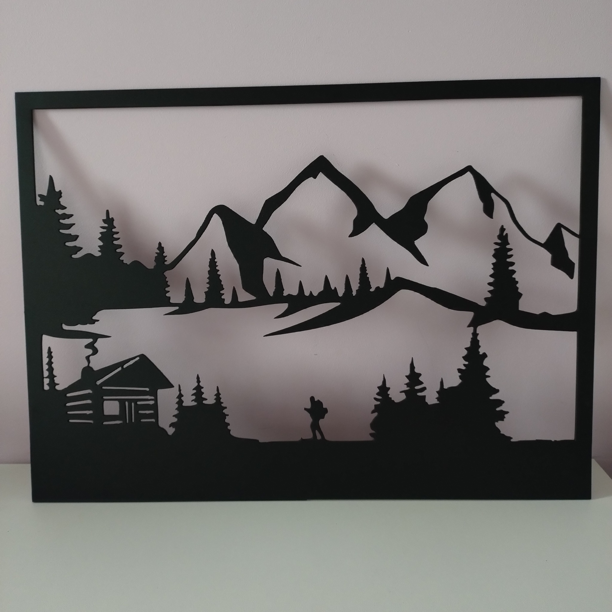 Czarna, metalowa dekoracja ścienna przedstawiająca górski krajobraz z chatką, drzewami iglastymi i sylwetką wędrowca, umieszczona w prostokątnej ramie.