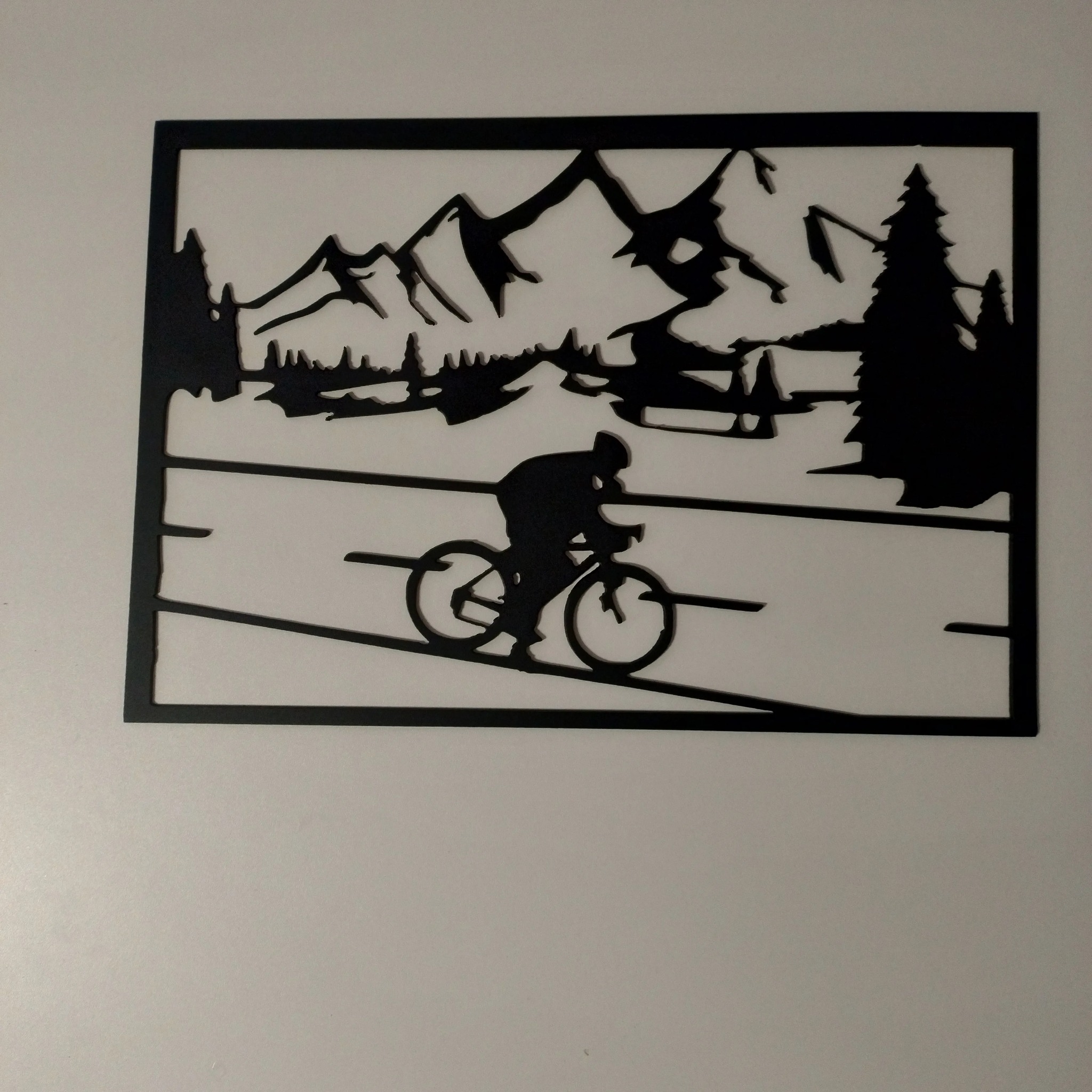 Czarna metalowa dekoracja ścienna przedstawiająca rowerzystę na tle górskiego krajobrazu z drzewami, wykonana prawdopodobnie techniką cięcia CNC.