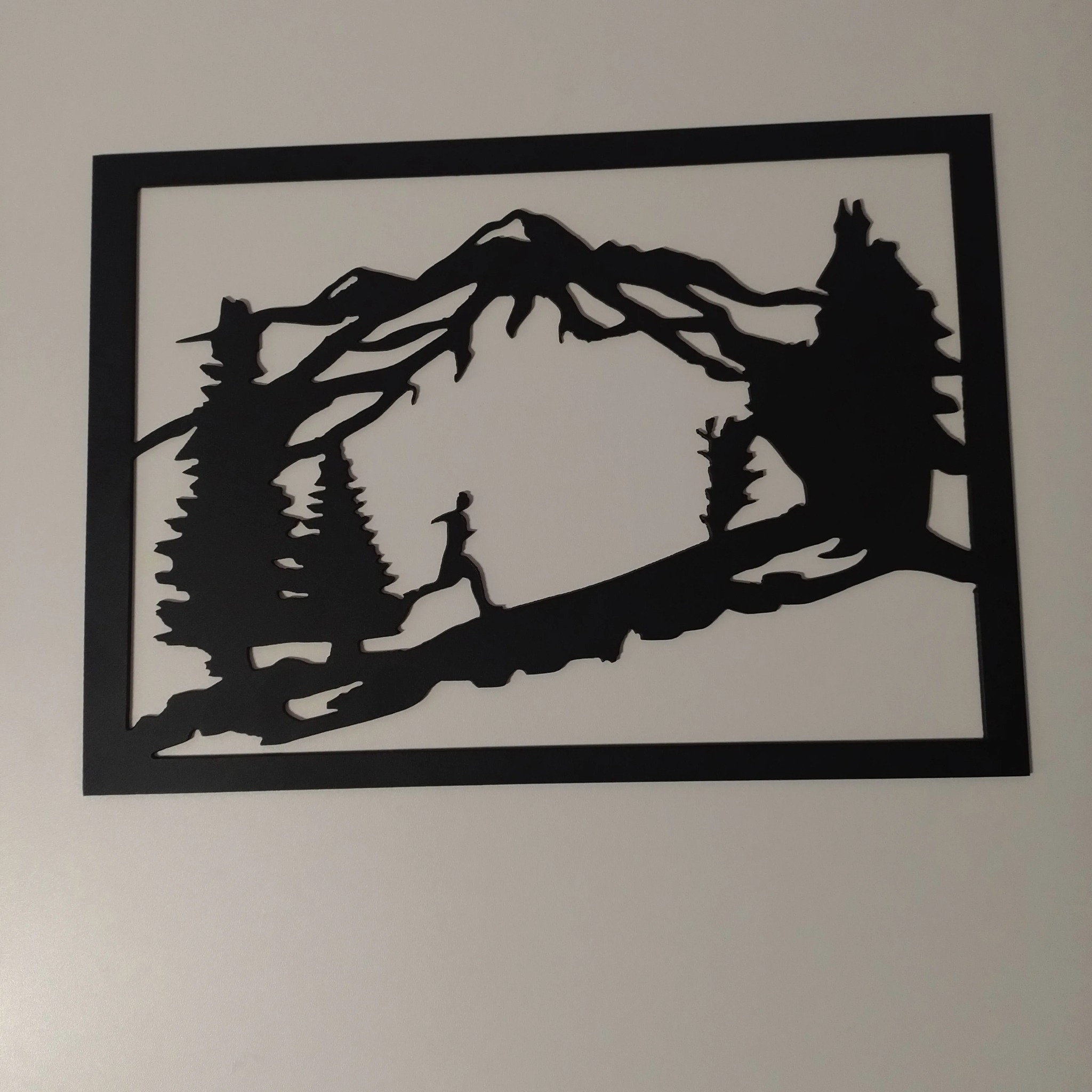 Czarna, laserowo wycinana dekoracja ścienna przedstawiająca sylwetkę biegacza w górskim krajobrazie z drzewami iglastymi, całość w prostokątnej ramie na jasnym tle ściany.