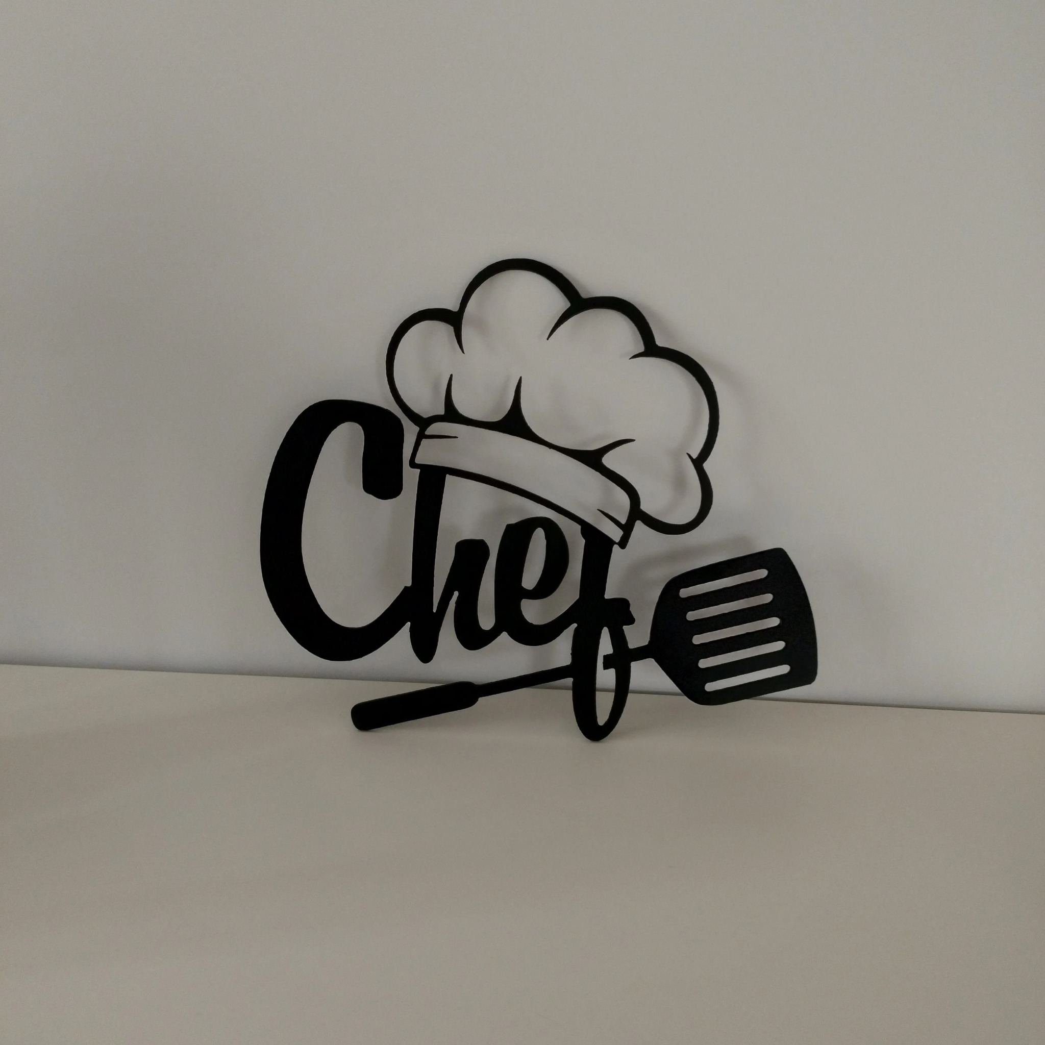 Czarny, wycięty laserowo napis 'Chef' z czapką kucharską i łopatką, umieszczony na jasnym tle.