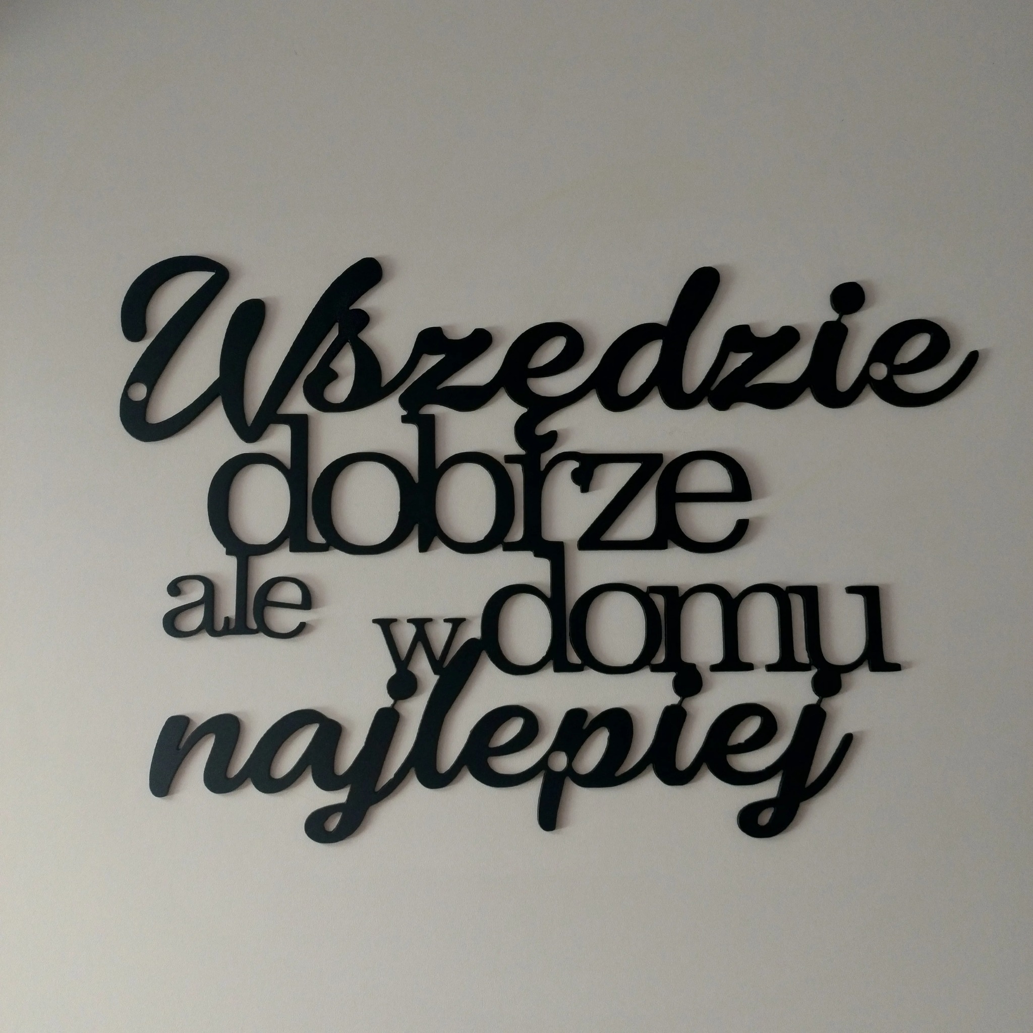 Dekoracja ścienna z wyciętymi laserowo czarnymi literami tworzącymi napis 'Wszędzie dobrze ale w domu najlepiej', przymocowana do jasnej ściany.