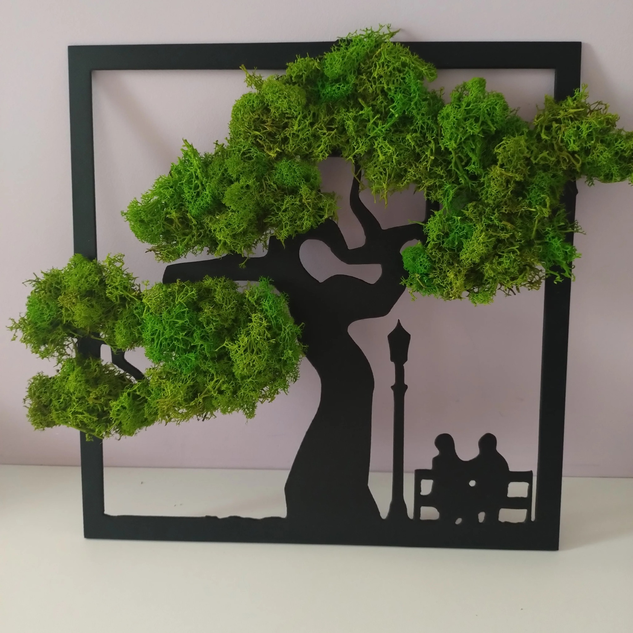 Dekoracja ścienna: czarna metalowa rama z sylwetką drzewa, lampy ulicznej i pary na ławce, korona drzewa wypełniona zielonym mchem.