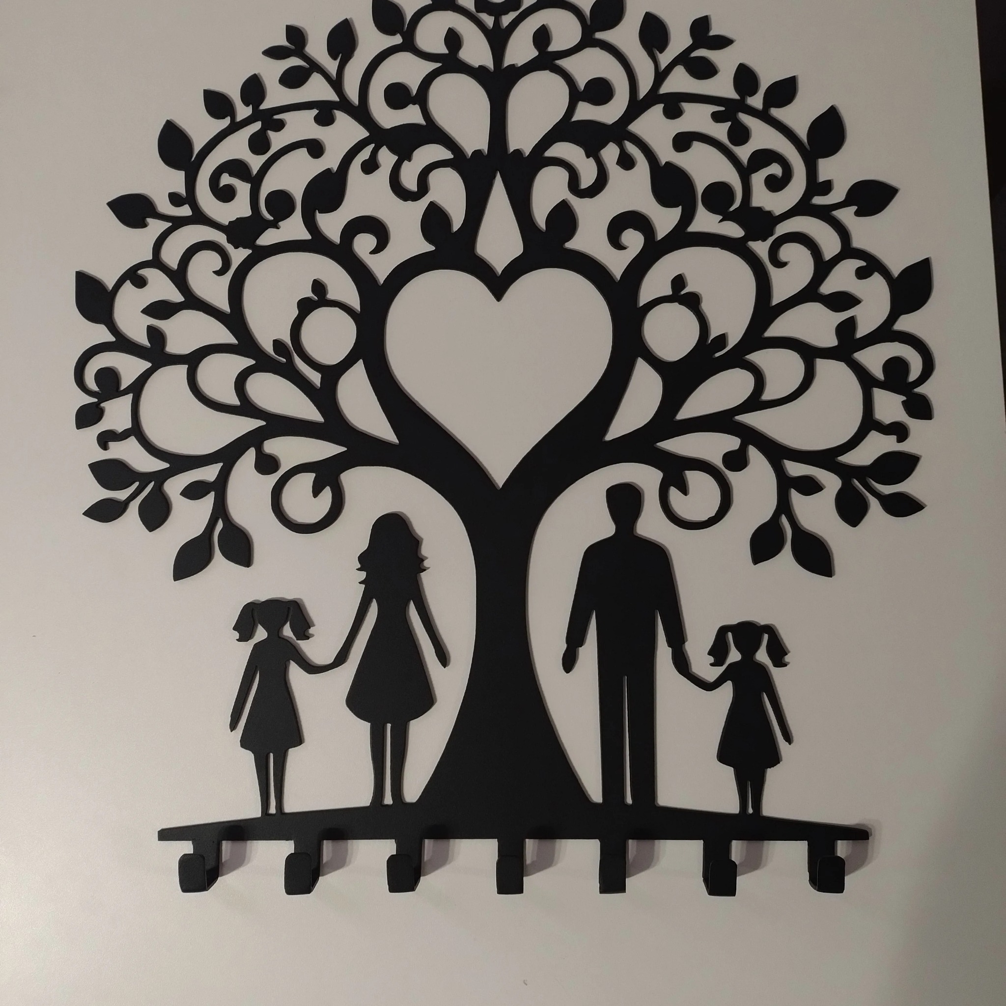 Czarny, metalowy dekor ścienny przedstawiający drzewo z sercem w koronie, pod którym stoją sylwetki rodziny: ojca, matki i dwóch córek. Dolna część drzewa pełni funkcję wieszaka z haczykami.