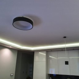 Bud - Nowoczesna kuchnia z podświetlanym sufitem LED i okrągłą lampą wiszącą, minimalistyczne białe szafki z połyskiem.