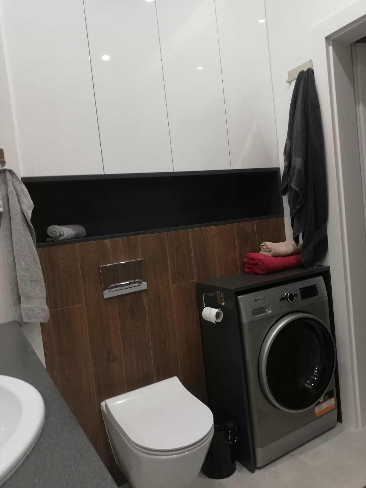 Nowoczesna łazienka z pralką obudowaną czarną szafką, wiszącą toaletą, brązowymi płytkami imitującymi drewno oraz białymi szafkami wiszącymi.