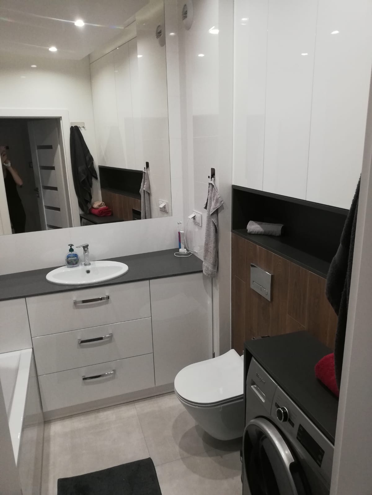 Nowoczesna łazienka z białymi szafkami, szarym blatem, umywalką, toaletą, pralką i dużym lustrem, w którym odbija się osoba robiąca zdjęcie.