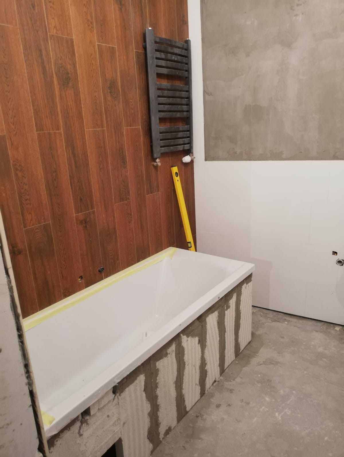 Biała wanna obudowana bloczkami i zaprawą, poziomica oparta o ścianę z brązowych paneli imitujących drewno, grzejnik drabinkowy na ścianie, etap prac w łazience.