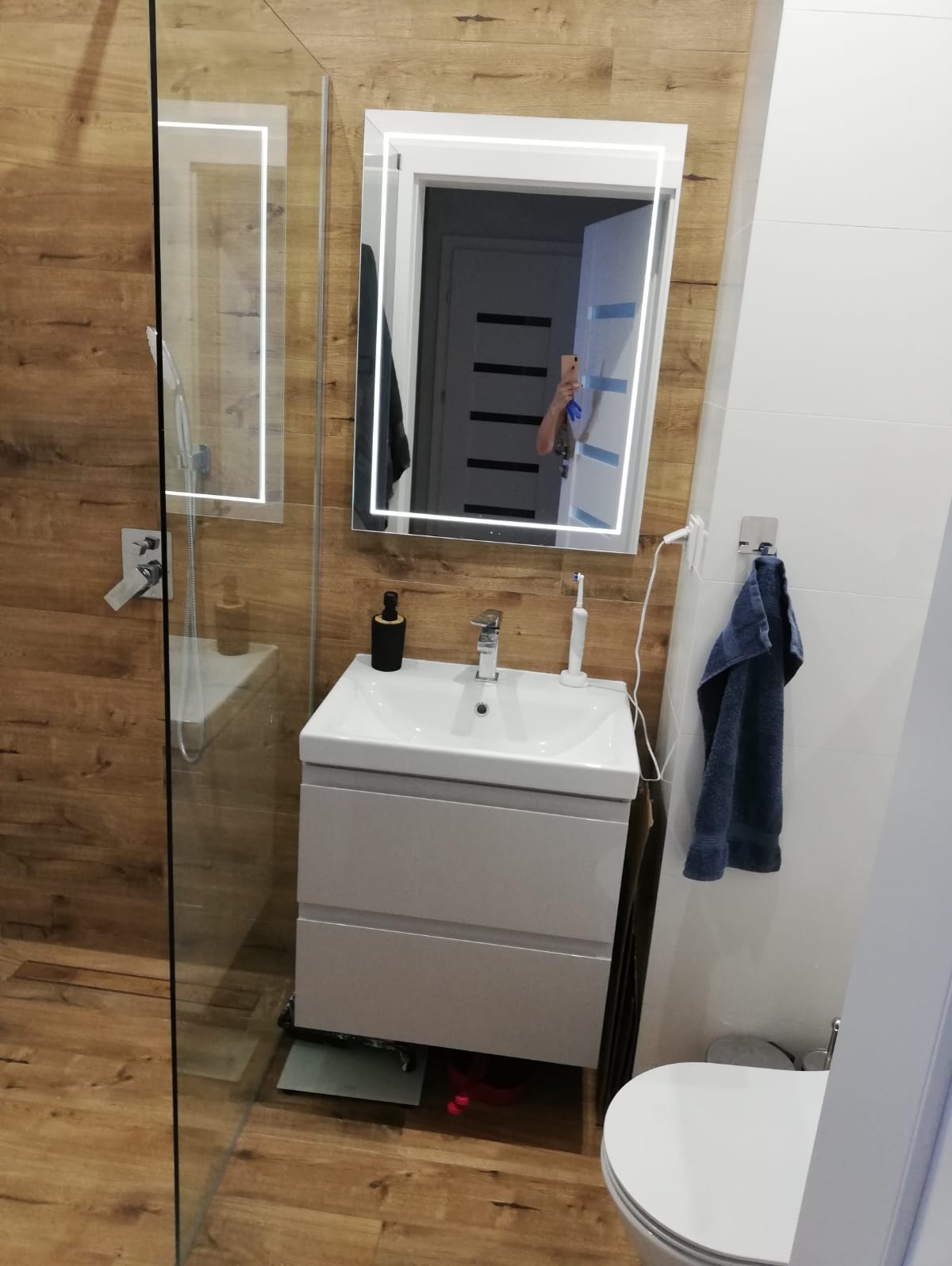 Nowoczesna łazienka z drewnopodobnymi płytkami, kabiną prysznicową ze szklaną ścianką, białą szafką umywalkową z lustrem podświetlanym LED, toaletą i niebieskim ręcznikiem.