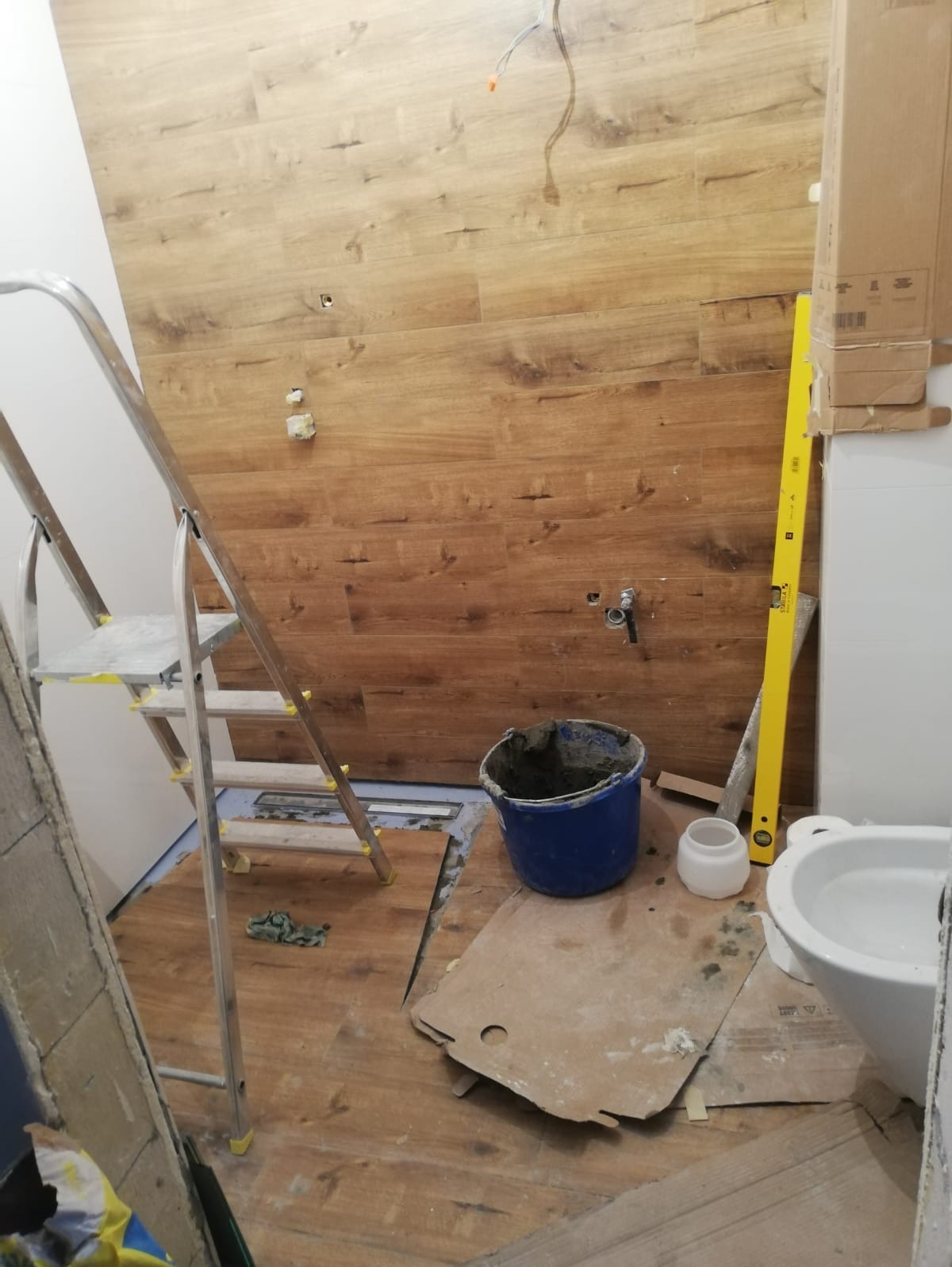 Łazienka w trakcie wykańczania: imitacja drewnianej okładziny ściennej, drabina, wiadro z zaprawą, poziomica i częściowo zamontowana armatura sanitarna.