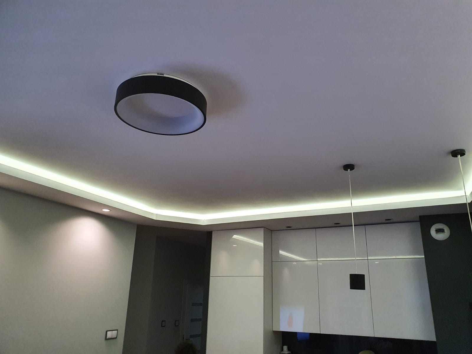 Nowoczesna kuchnia z podświetlanym sufitem LED i okrągłą lampą wiszącą, minimalistyczne białe szafki z połyskiem.