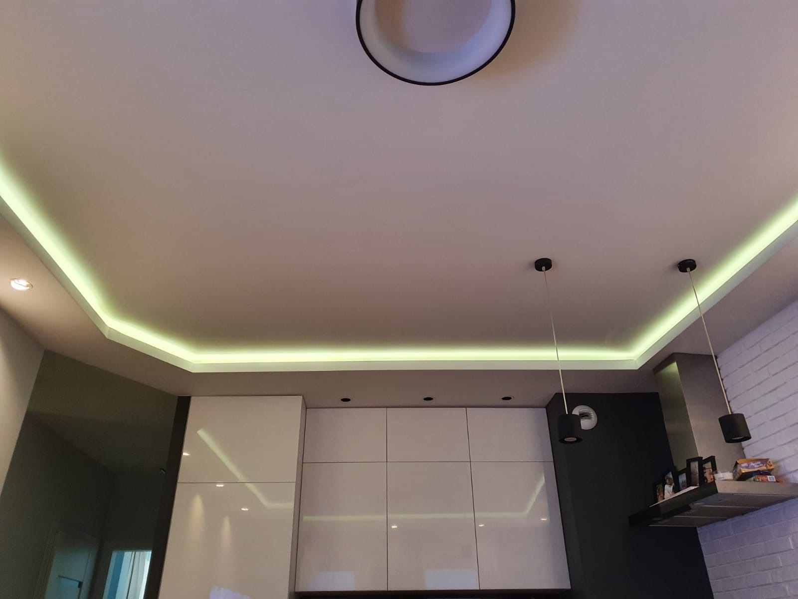 Nowoczesna kuchnia w Olsztynie z białymi szafkami i podświetlanym sufitem wielopoziomowym z oświetleniem LED, widoczne również wiszące lampy i ceglana ściana.