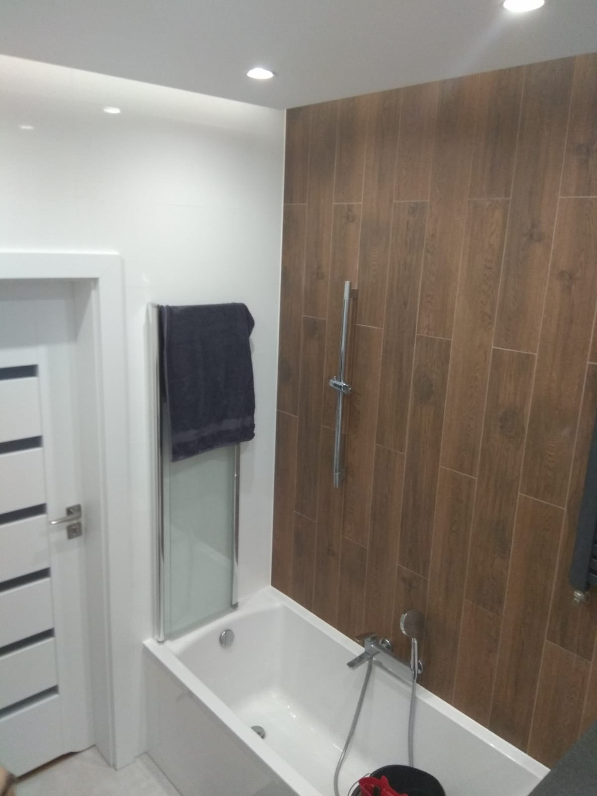 Nowoczesna łazienka z białą wanną, szklanym parawanem nawannowym, ręcznikiem na grzejniku i ścianą wyłożoną brązowymi płytkami imitującymi drewno.