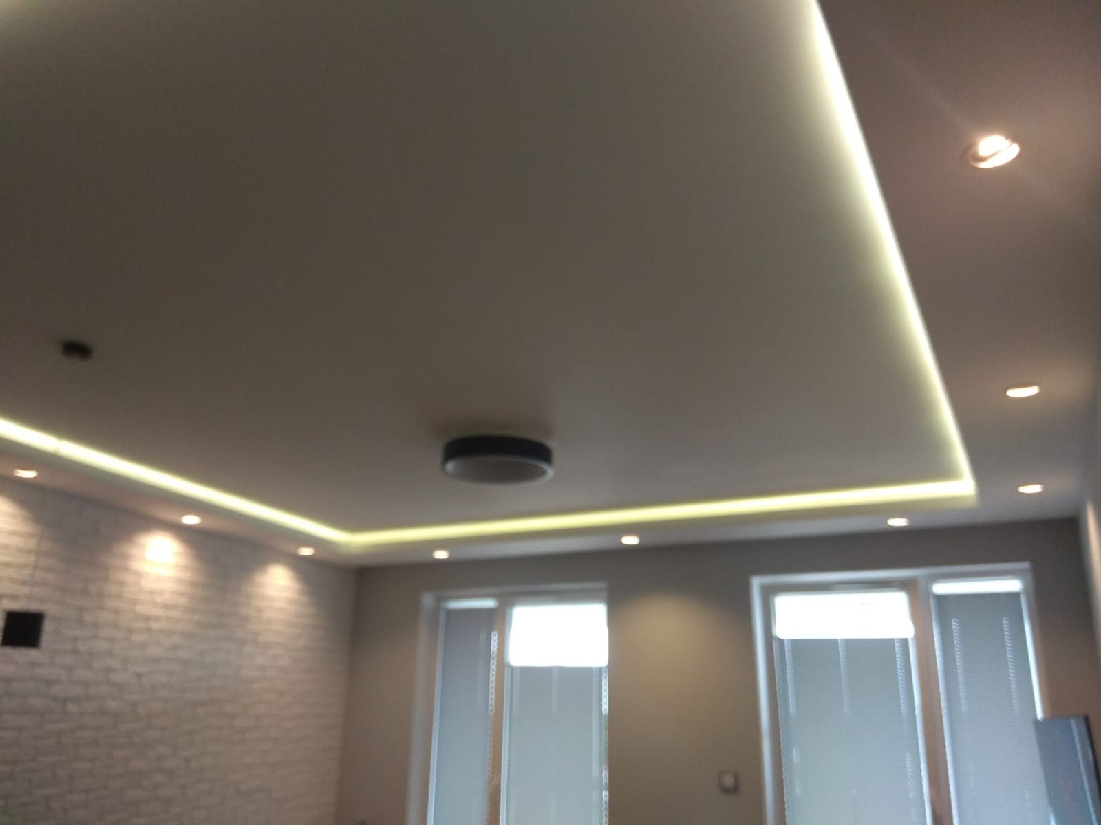 Nowoczesny sufit podwieszany z oświetleniem LED wzdłuż krawędzi, centralna okrągła lampa sufitowa, ściana z białej cegły w tle, widoczne okna z żaluzjami.